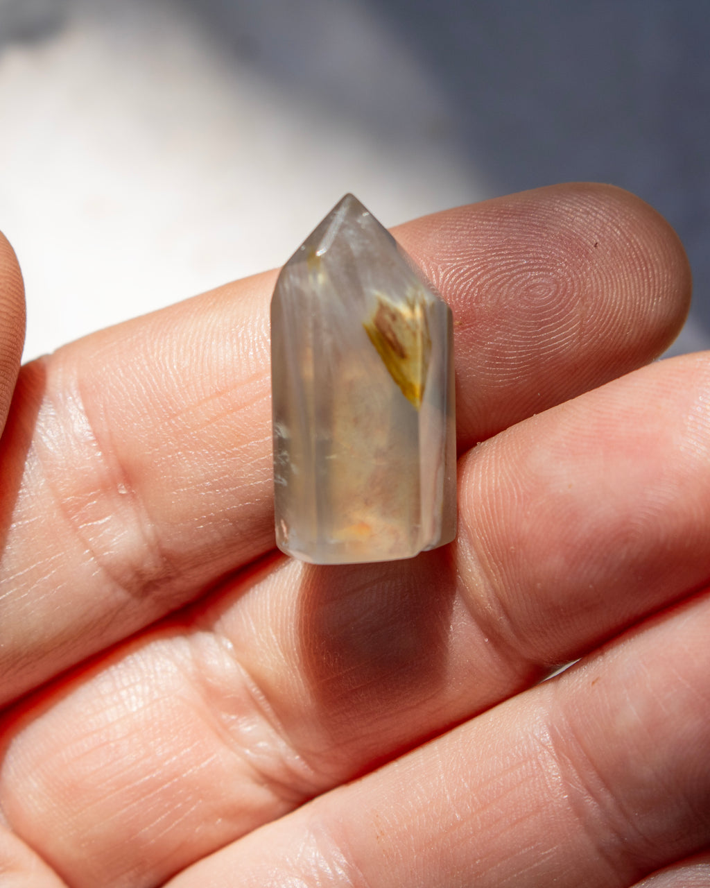 A small pointed clear crystal or gemstone held between fingers. The stone appears translucent milky white with a small yellow-brown inclusions or feather-like feature inside.
