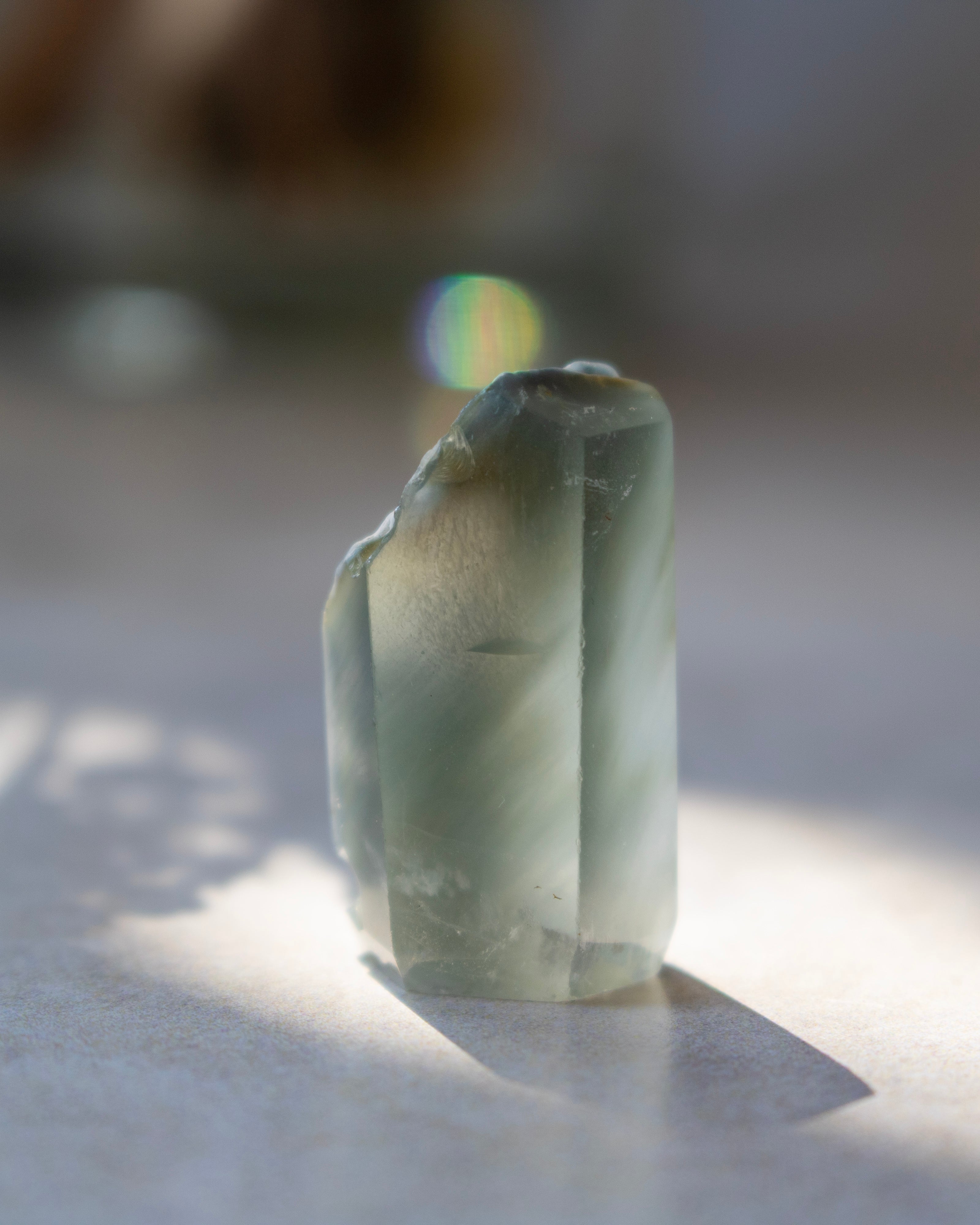 A natural pointed crystal stone with translucent milky white and warm brown inclusions, standing upright on a light surface under direct sunlight with a soft shadow.