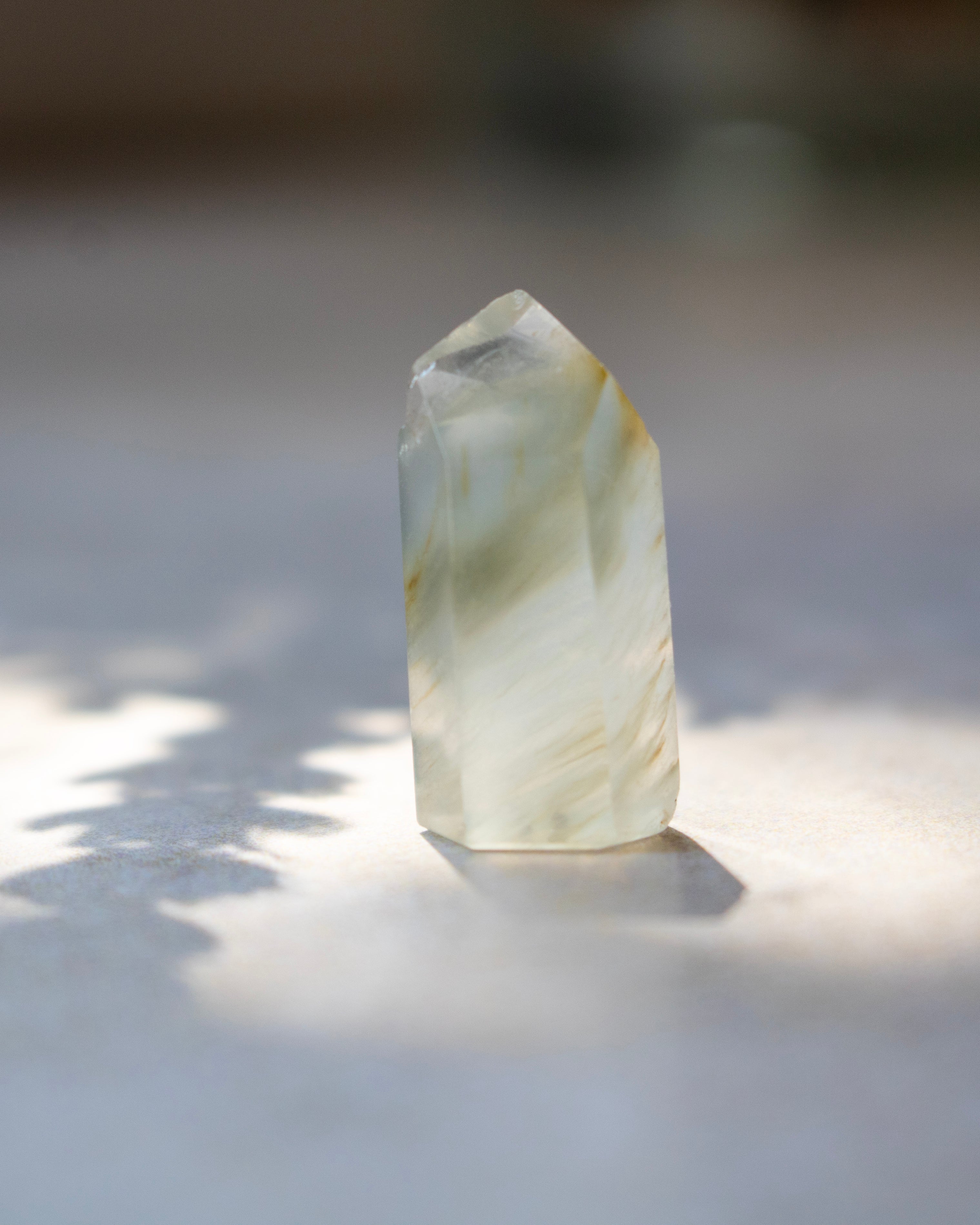 A natural pointed crystal stone with white, milky translucent bands and soft yellow-brown veining, standing upright on a light surface with dappled sunlight and shadows.