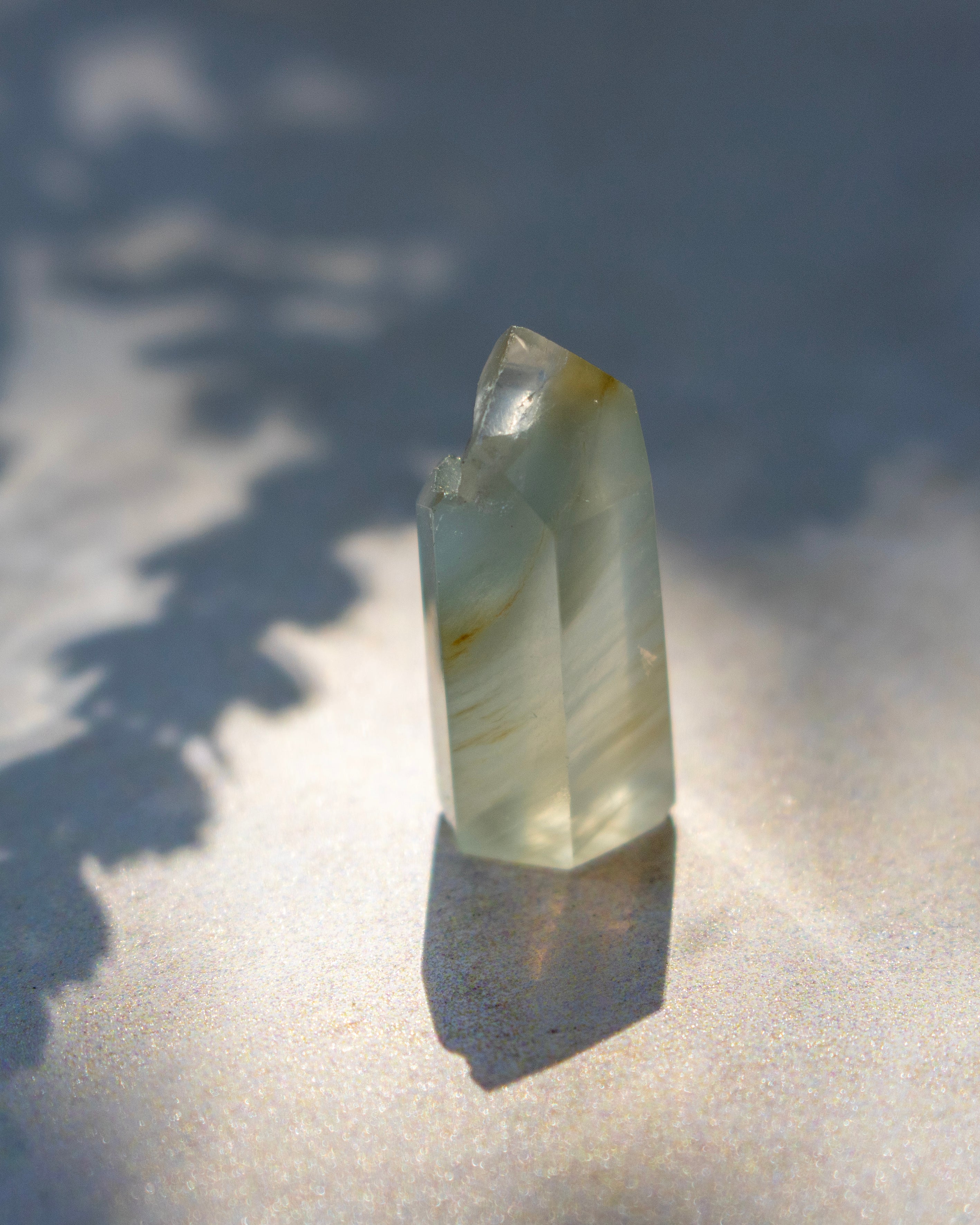 A natural translucent crystal point with pale green and white marbling, standing upright on a sandy surface under direct sunlight with leaf shadows.