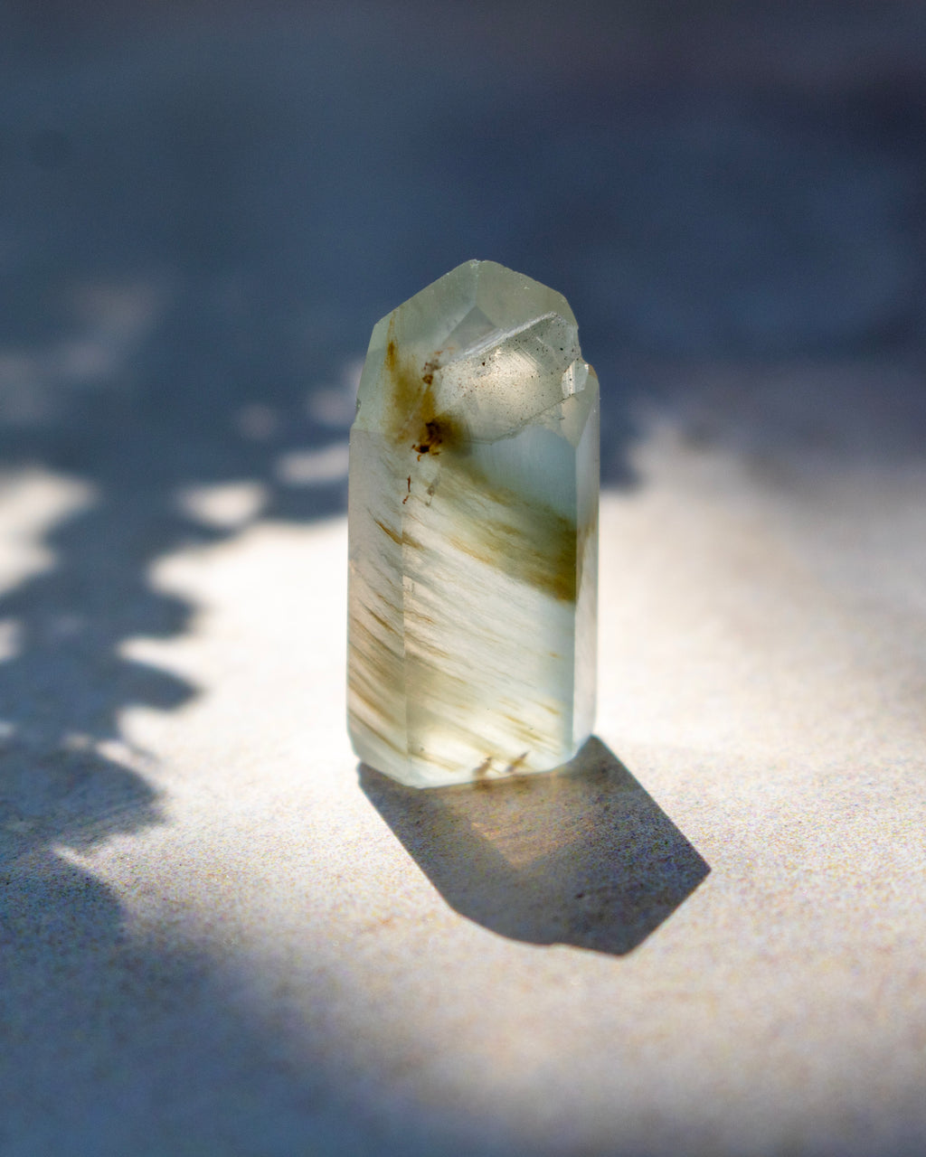 A natural clear quartz crystal point with internal inclusions and brownish streaks, standing upright on a textured surface under direct sunlight with a sharp shadow.