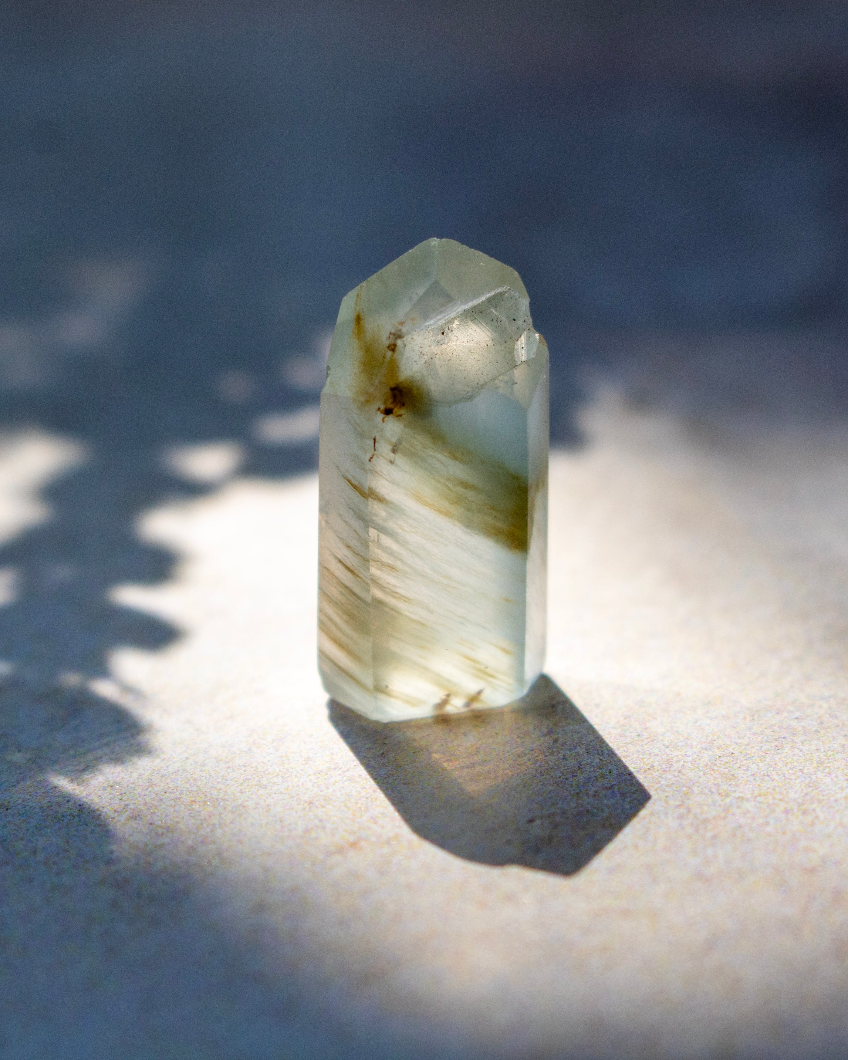 A natural clear quartz crystal point with internal inclusions and brownish streaks, standing upright on a textured surface under direct sunlight with a sharp shadow.