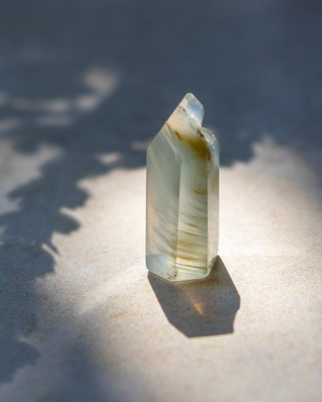 A natural clear quartz crystal point with subtle brown and yellow inclusions, standing upright on a textured beige surface under direct sunlight with a sharp shadow.