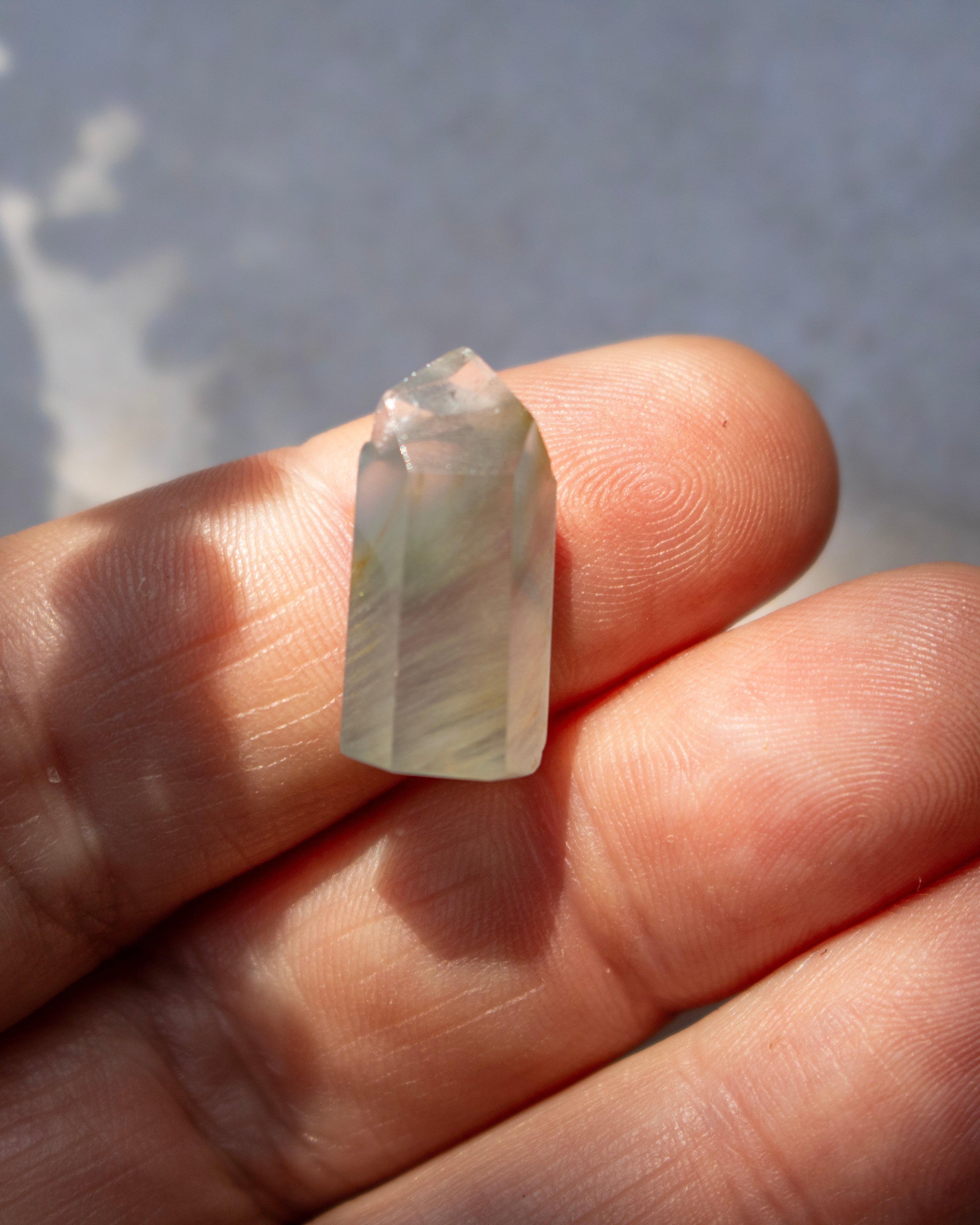 A small translucent pale green crystal point resting on a person's fingers against a soft gray background.