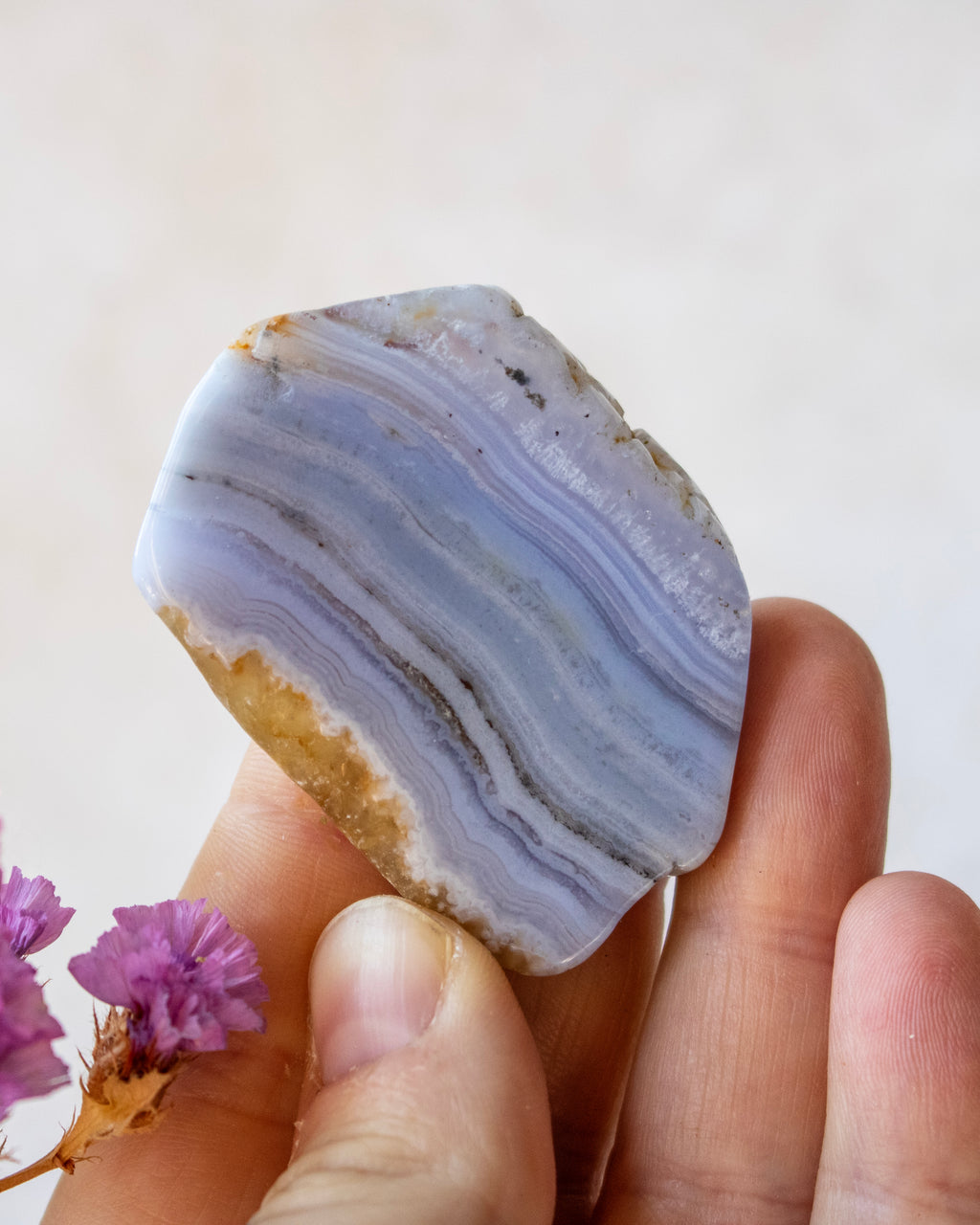 A hand holding a polished agate stone with lavender and light blue banded patterns, with small purple flowers in the corner against a soft beige background.