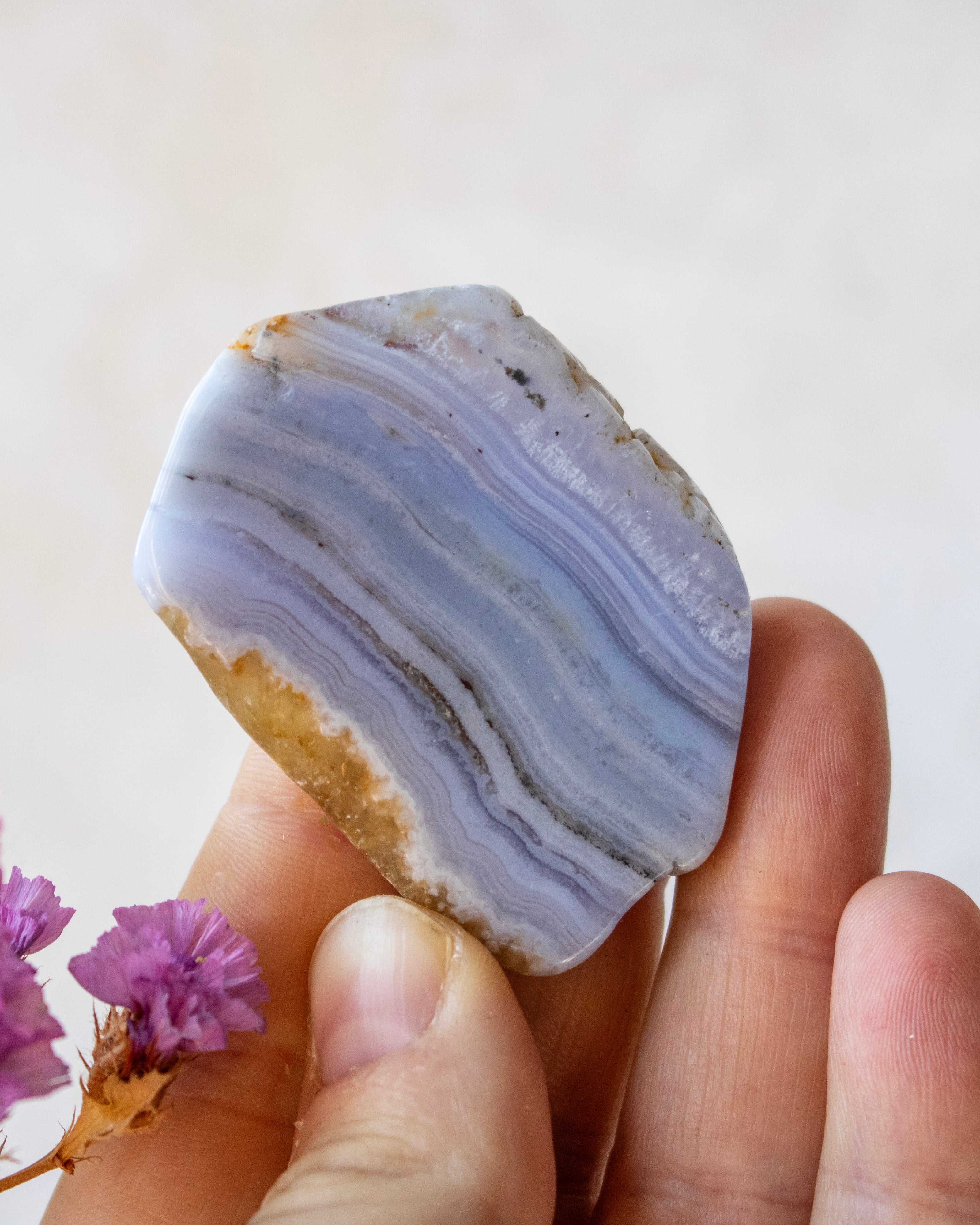 A hand holding a polished agate stone with lavender and light blue banded patterns, with small purple flowers in the corner against a soft beige background.