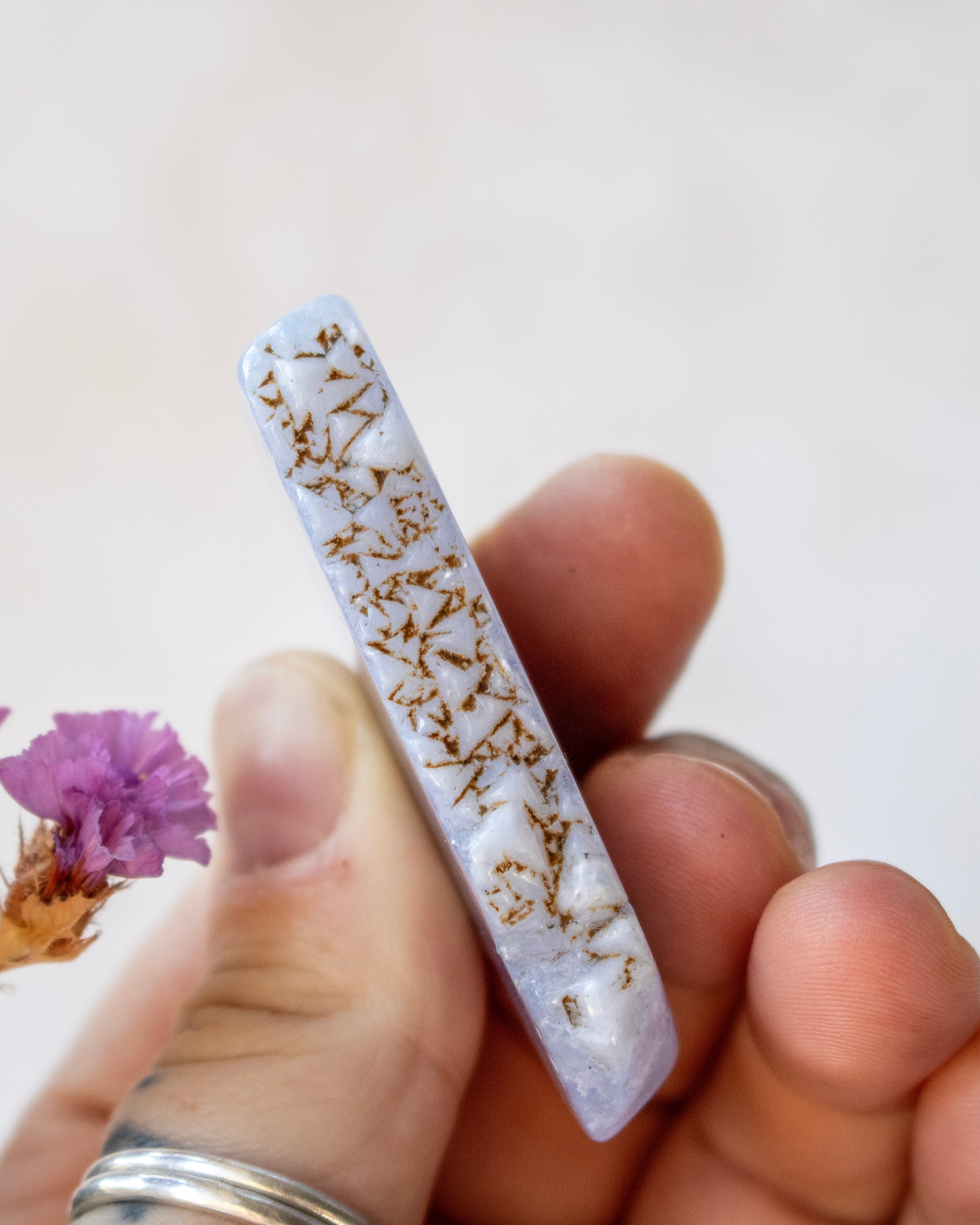 A hand holding a polished rectangular stone or crystal with a pale lavender-gray body and dense metallic gold branching inlay, against a soft light background with a small purple flower at the side.