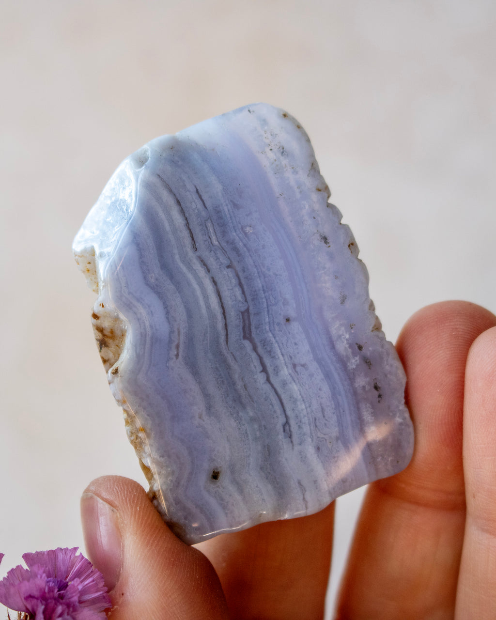 A hand holding a polished natural agate stone with layered blue, lavender, and white bands, set against a soft beige background.