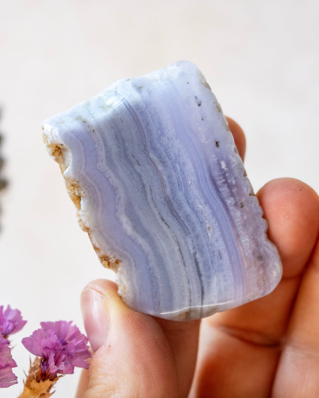 A hand holding a polished rectangular piece of lavender agate crystal with natural banding and rough mineral edges, against a soft light beige background with small dried flowers at the lower left.