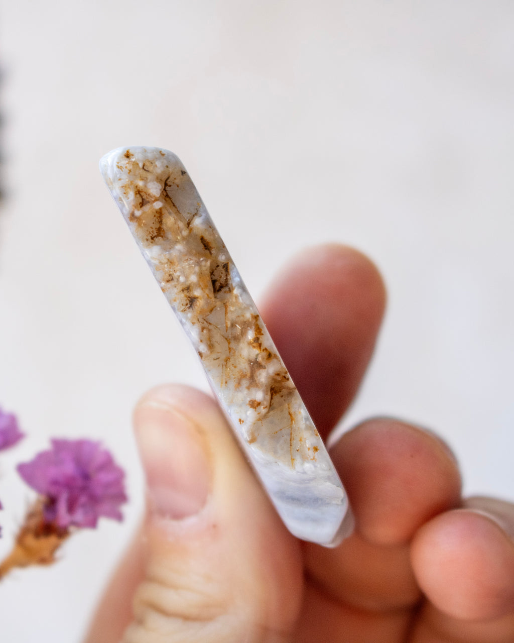 A hand holding a polished white crystal point with prominent brown and gold matrix inclusions, set against a soft, out-of-focus light background with small purple flowers in the corner.