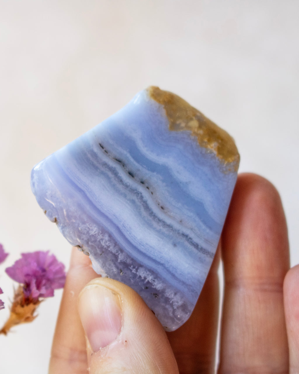 A hand holding a polished natural stone with layered light blue, lavender, and white bands, featuring a rough tan textured top edge, against a soft beige background with small dried flowers at the side.
