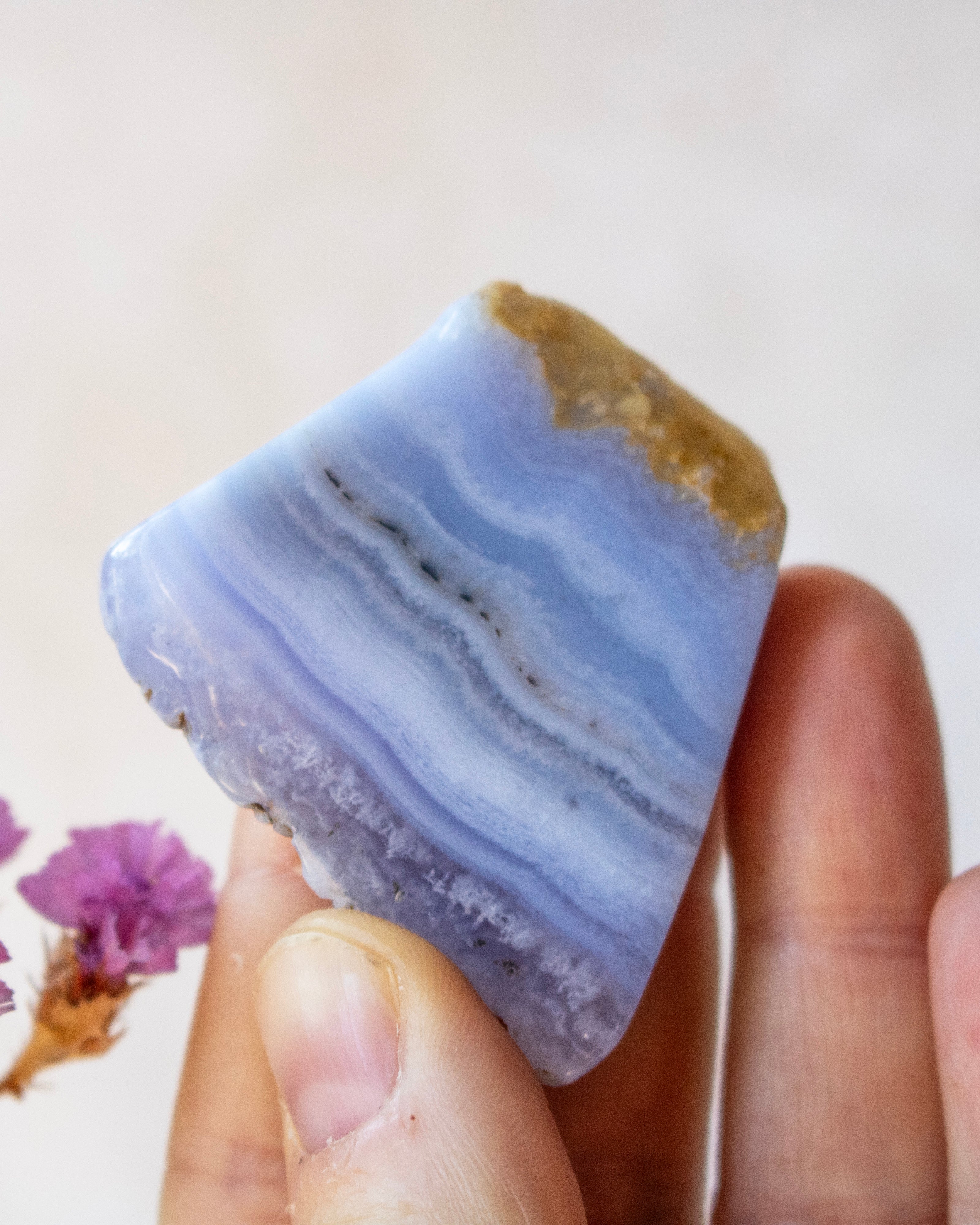 A hand holding a polished natural stone with layered light blue, lavender, and white bands, featuring a rough tan textured top edge, against a soft beige background with small dried flowers at the side.