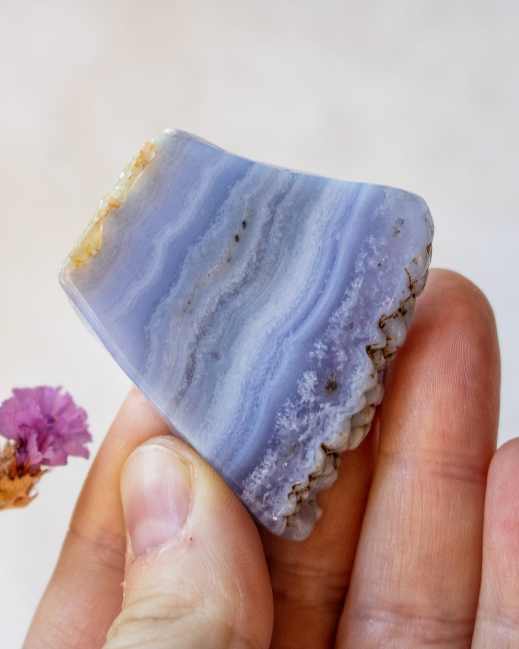 A hand holding a polished lavender-blue agate crystal slice with natural geode formations and a rough brown outer edge, against a soft beige background.
