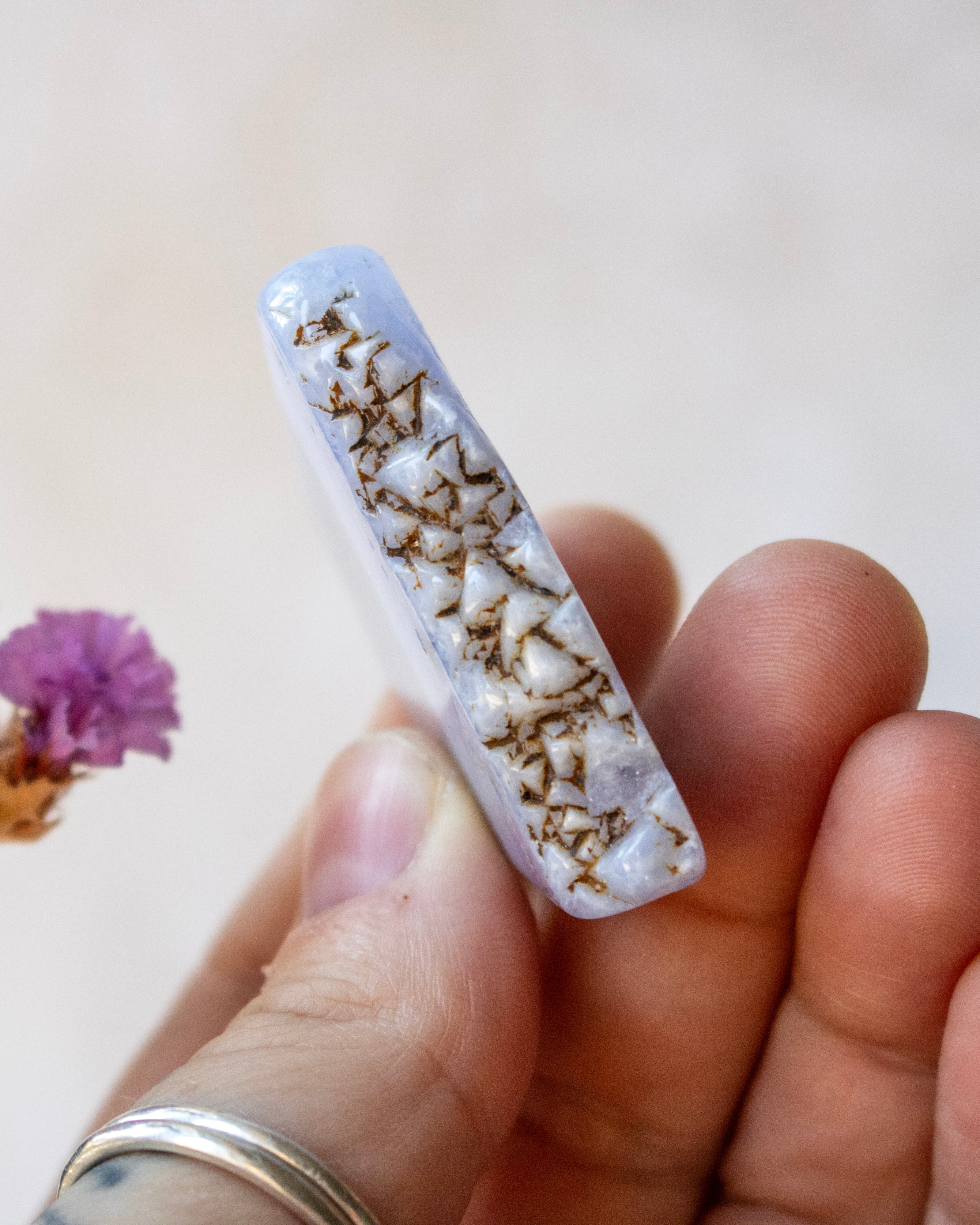 A hand holding a polished white crystal point with brown and gold mineral inclusions, against a soft beige background with a small dried purple flower on the left.
