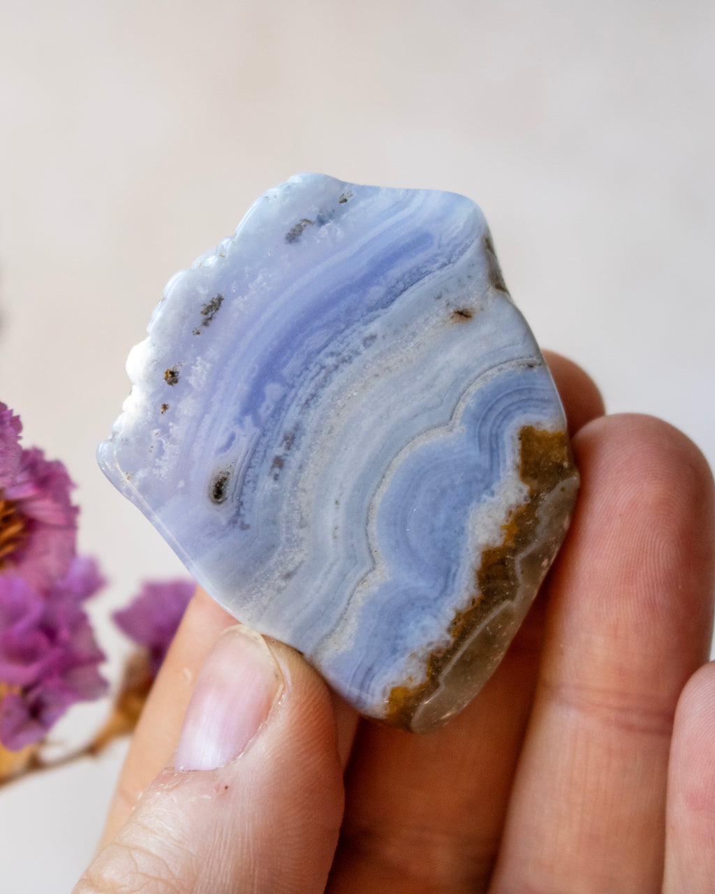 A hand holding a polished natural agate crystal with layered blue, white, and brown banding patterns against a soft beige background.