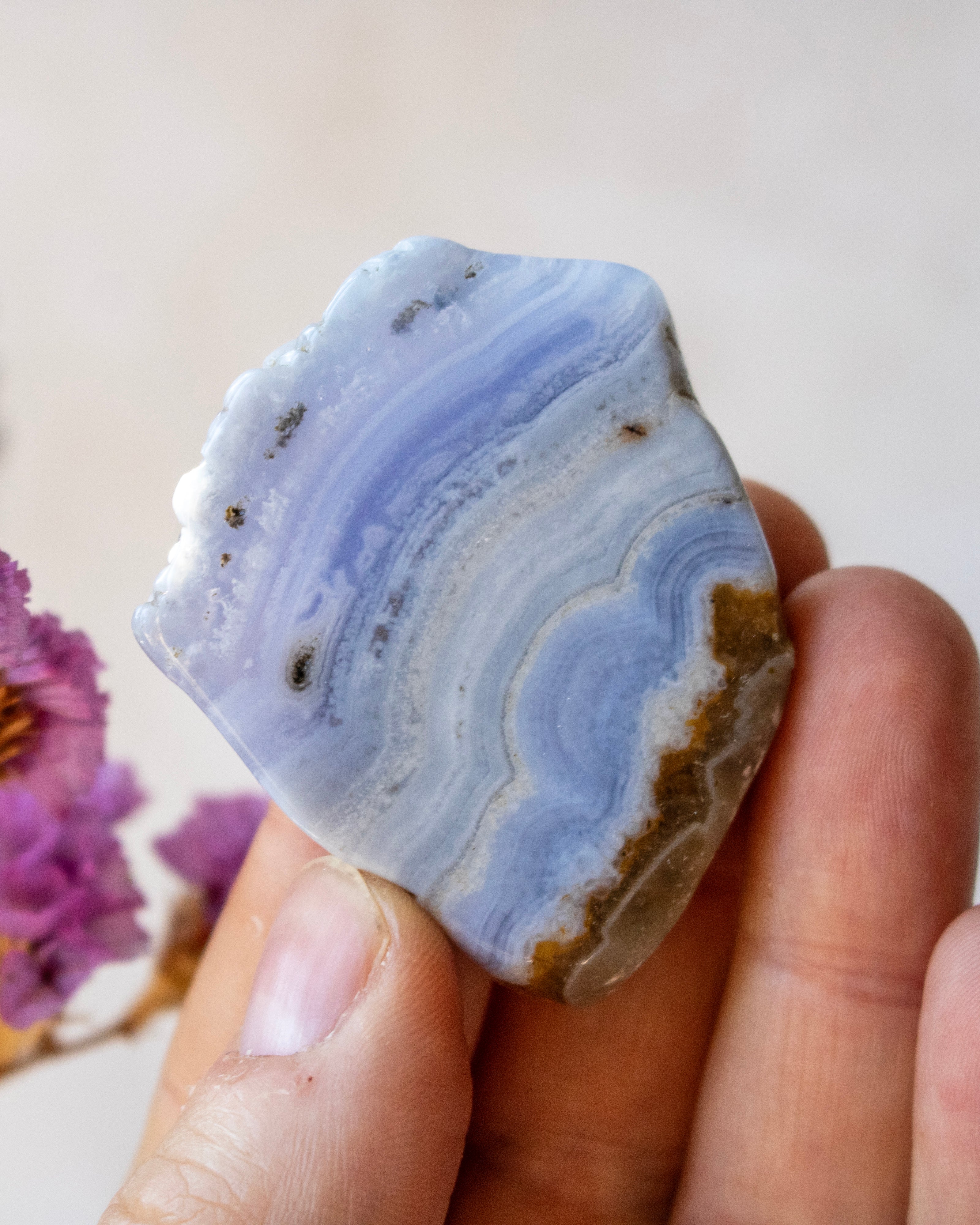 A hand holding a polished natural agate crystal with layered blue, white, and brown banding patterns against a soft beige background.