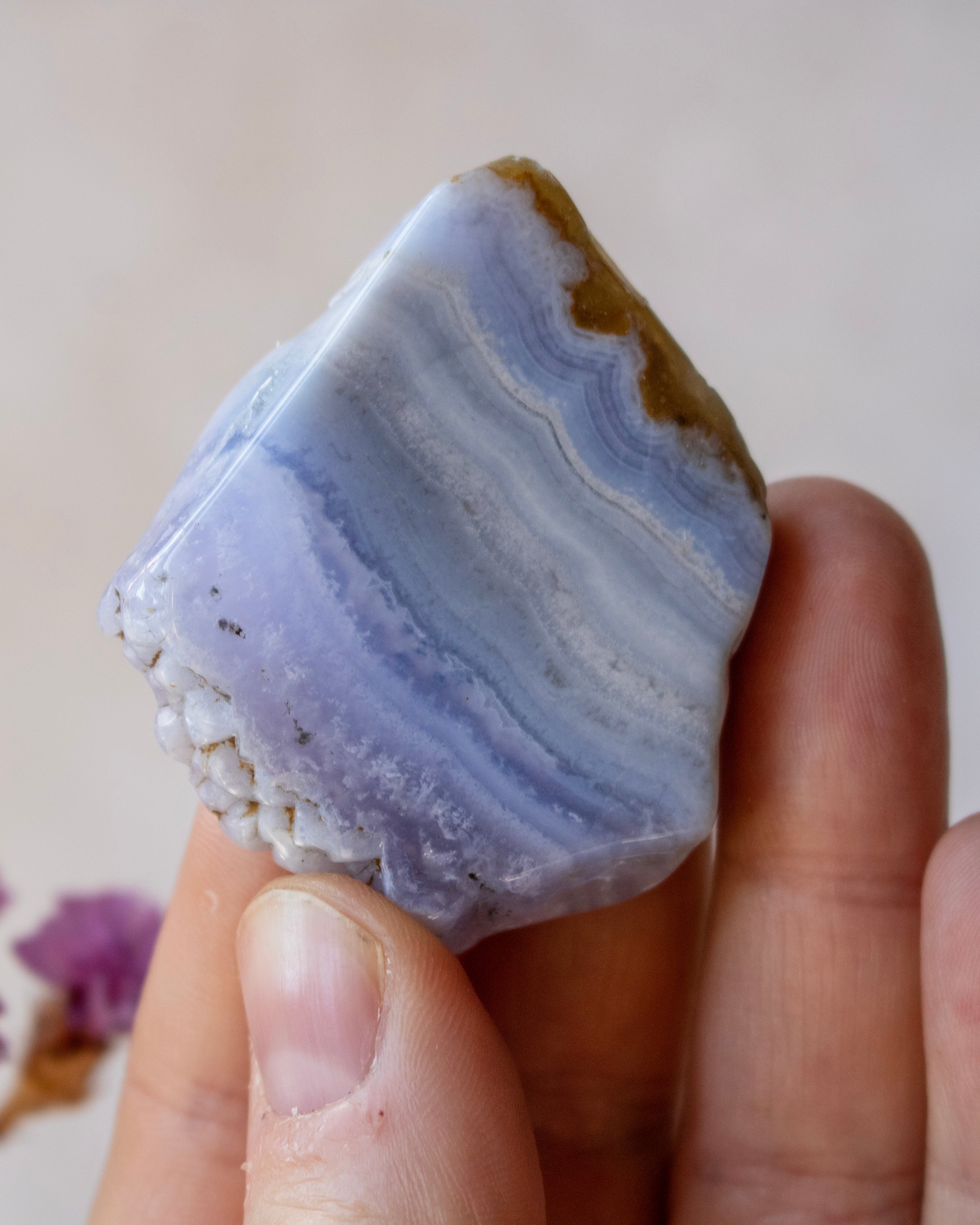 A hand holding a polished natural agate crystal with layered bands of light blue, lavender, white, and a brown rough edge, against a soft beige background.
