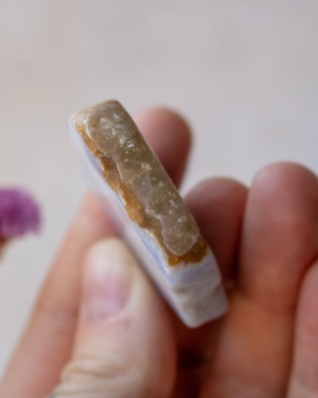 A hand holding a small rectangular stone or crystal slab with a rough tan-brown outer layer and a pale lavender-white inner face, shown close up against a soft light background.