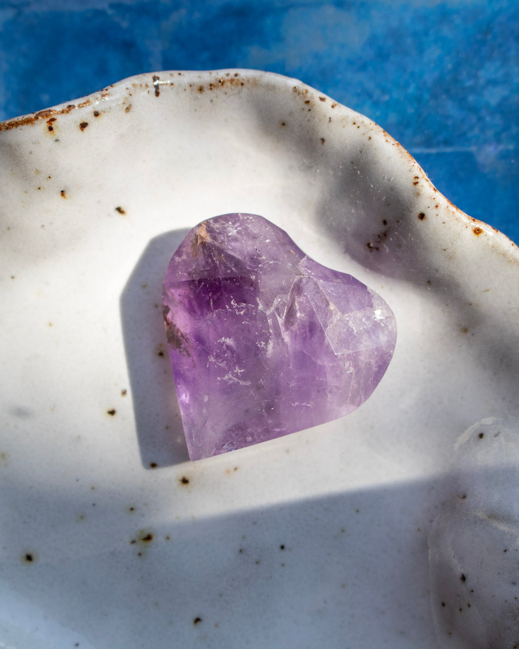 A heart-shaped purple amethyst crystal resting in a white ceramic dish with brown speckles, set against a textured blue background.