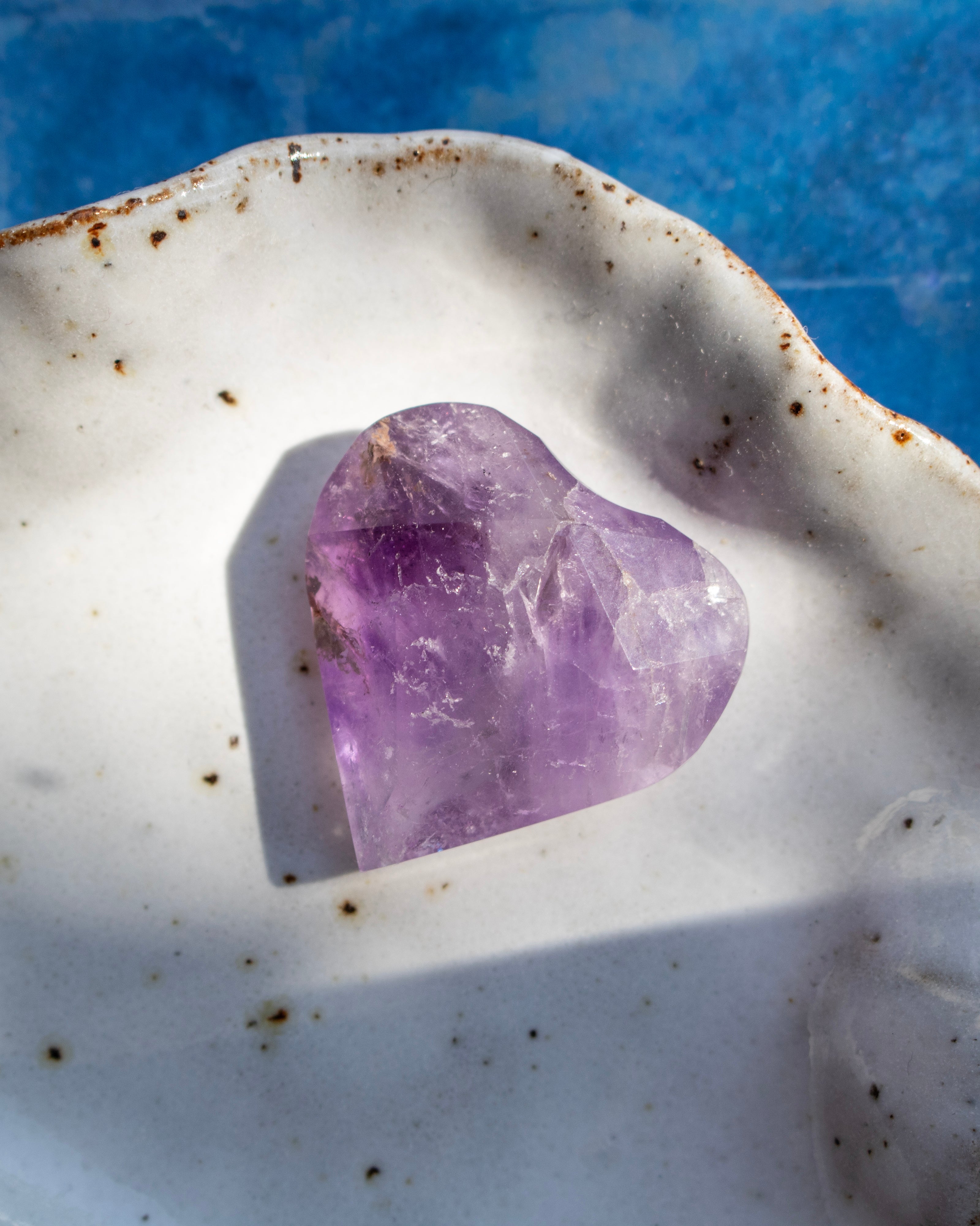 A heart-shaped purple amethyst crystal resting in a white ceramic dish with brown speckles, set against a textured blue background.