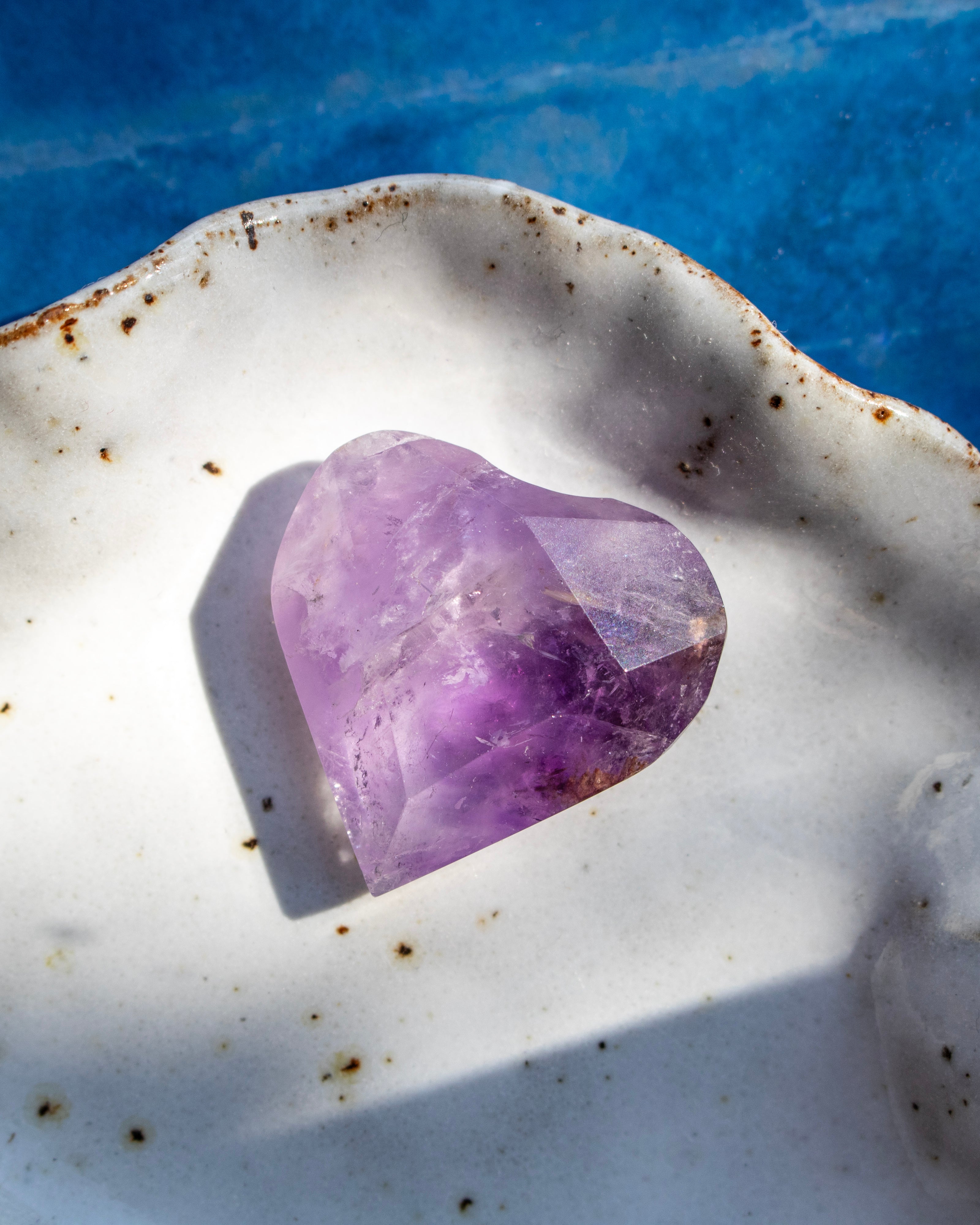 A heart-shaped purple amethyst crystal resting on a white ceramic dish with brown speckles, set against a bright blue background.
