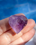A faceted heart-shaped purple crystal gemstone held in a person's hand against a blurred blue background.