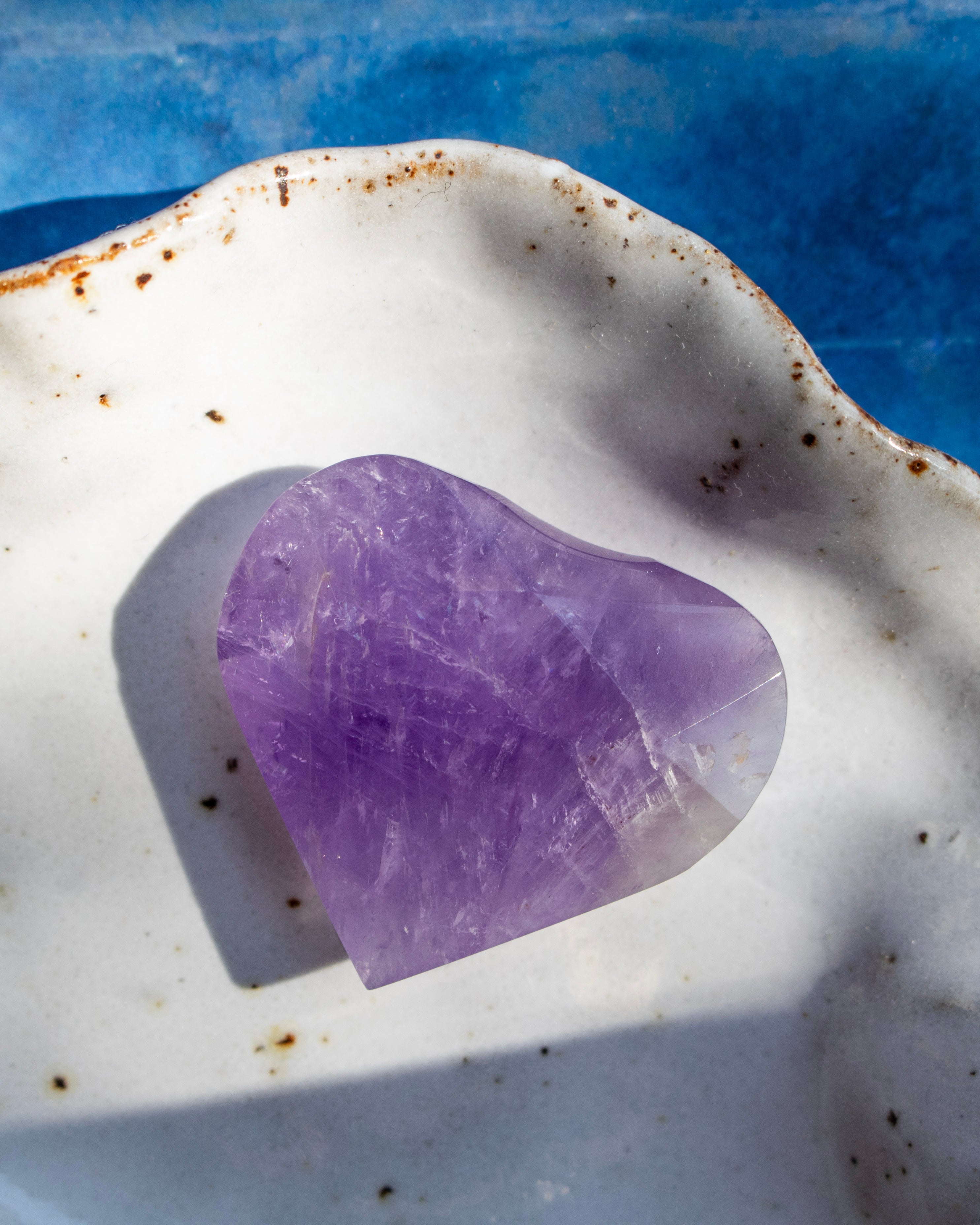 A polished purple amethyst heart-shaped crystal resting on a white speckled ceramic dish with a blue background.