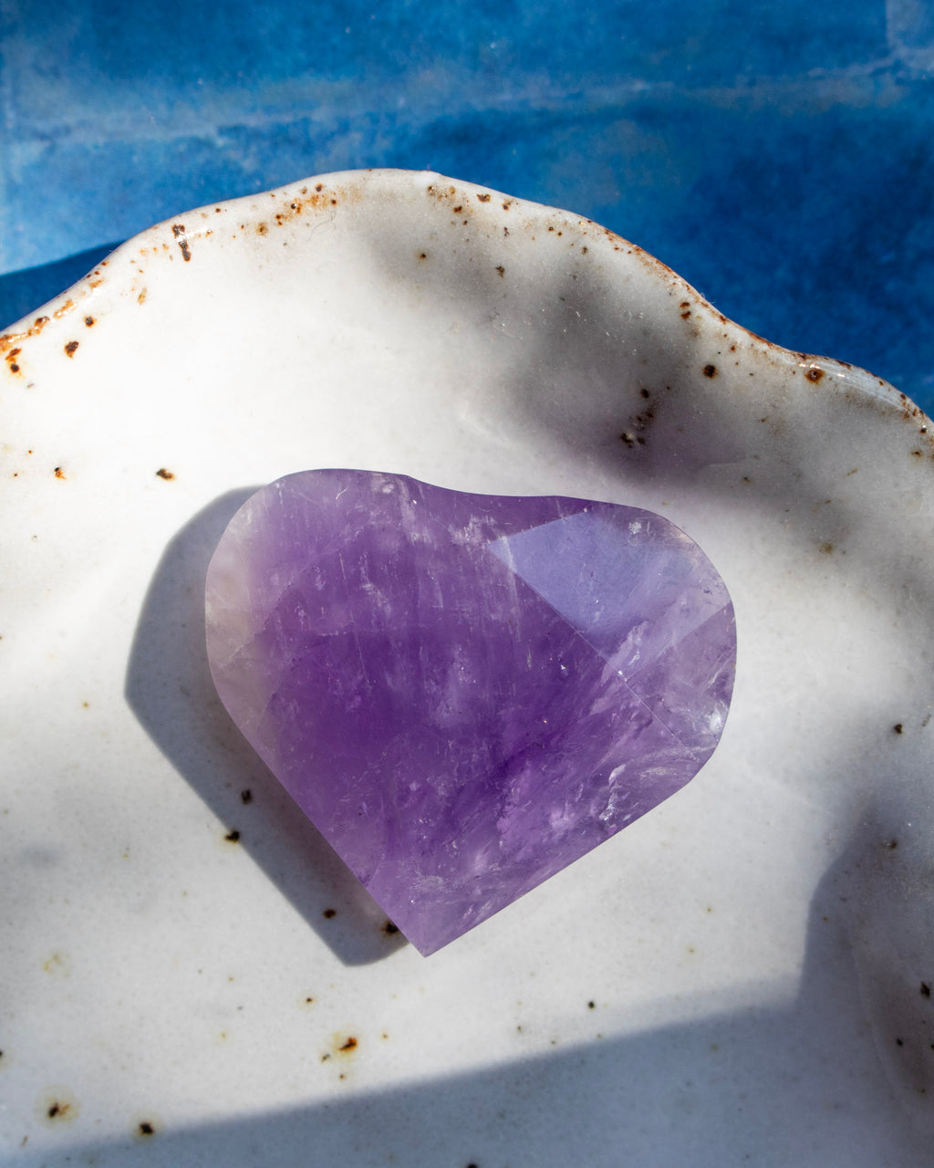 A heart-shaped purple amethyst crystal resting on a white speckled ceramic dish with a blue background.
