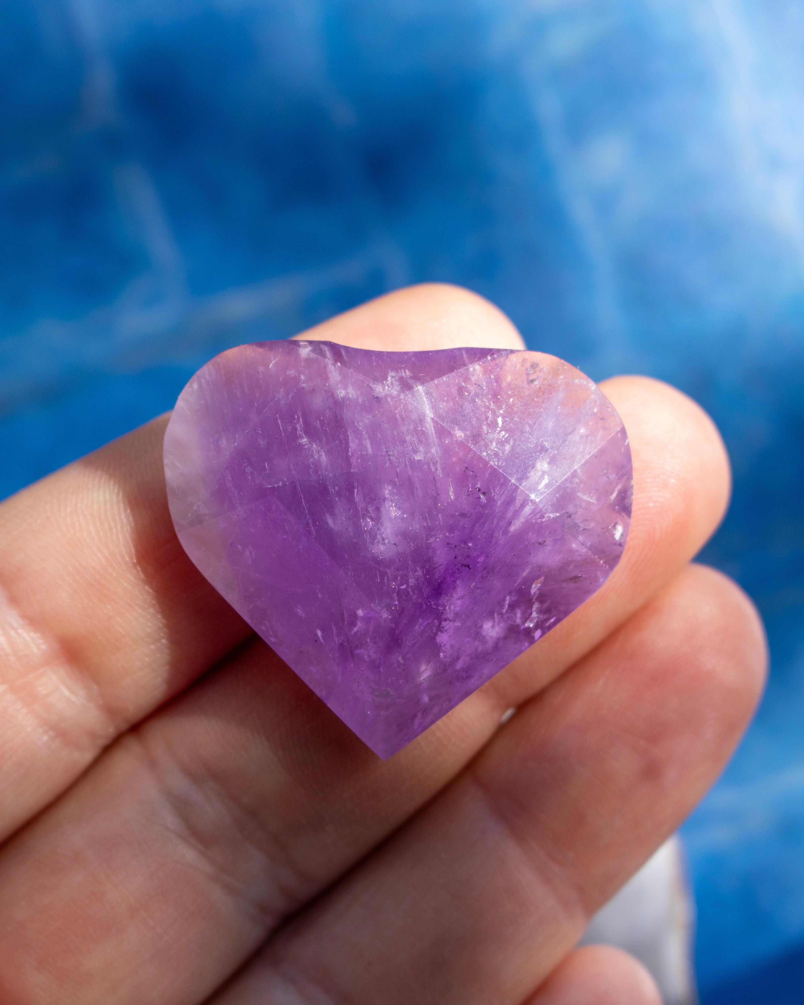 A heart-shaped purple amethyst crystal held in a person's hand against a blurred blue background.