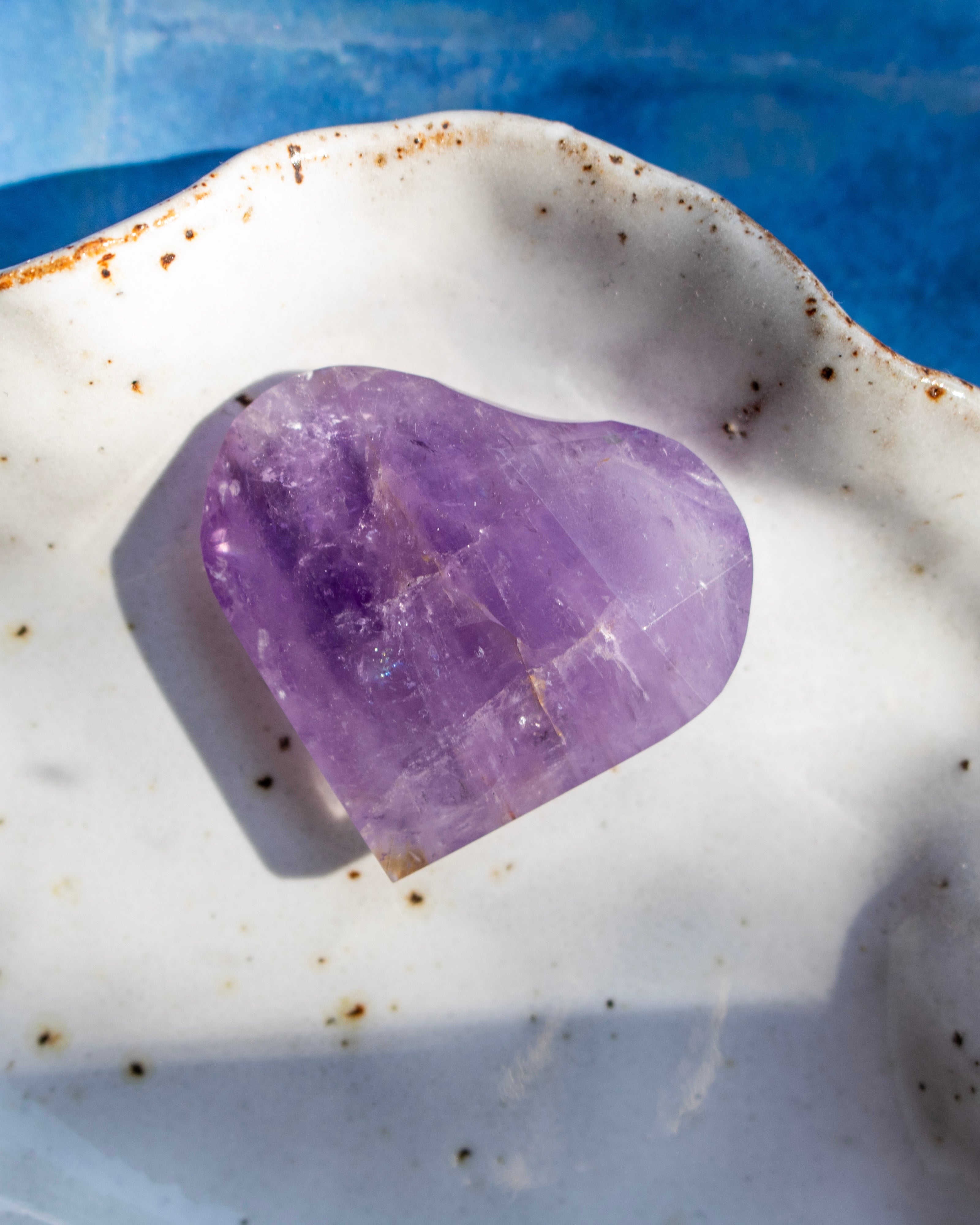 A heart-shaped purple amethyst crystal resting on a white ceramic dish with brown speckles, set against a blue background.