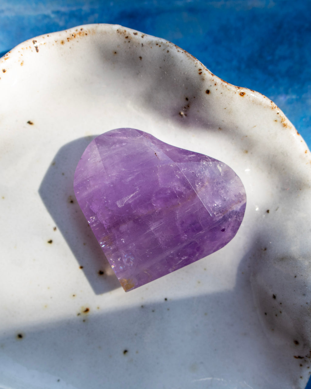 A heart-shaped purple crystal gemstone resting on a white ceramic dish with a blue textured background.