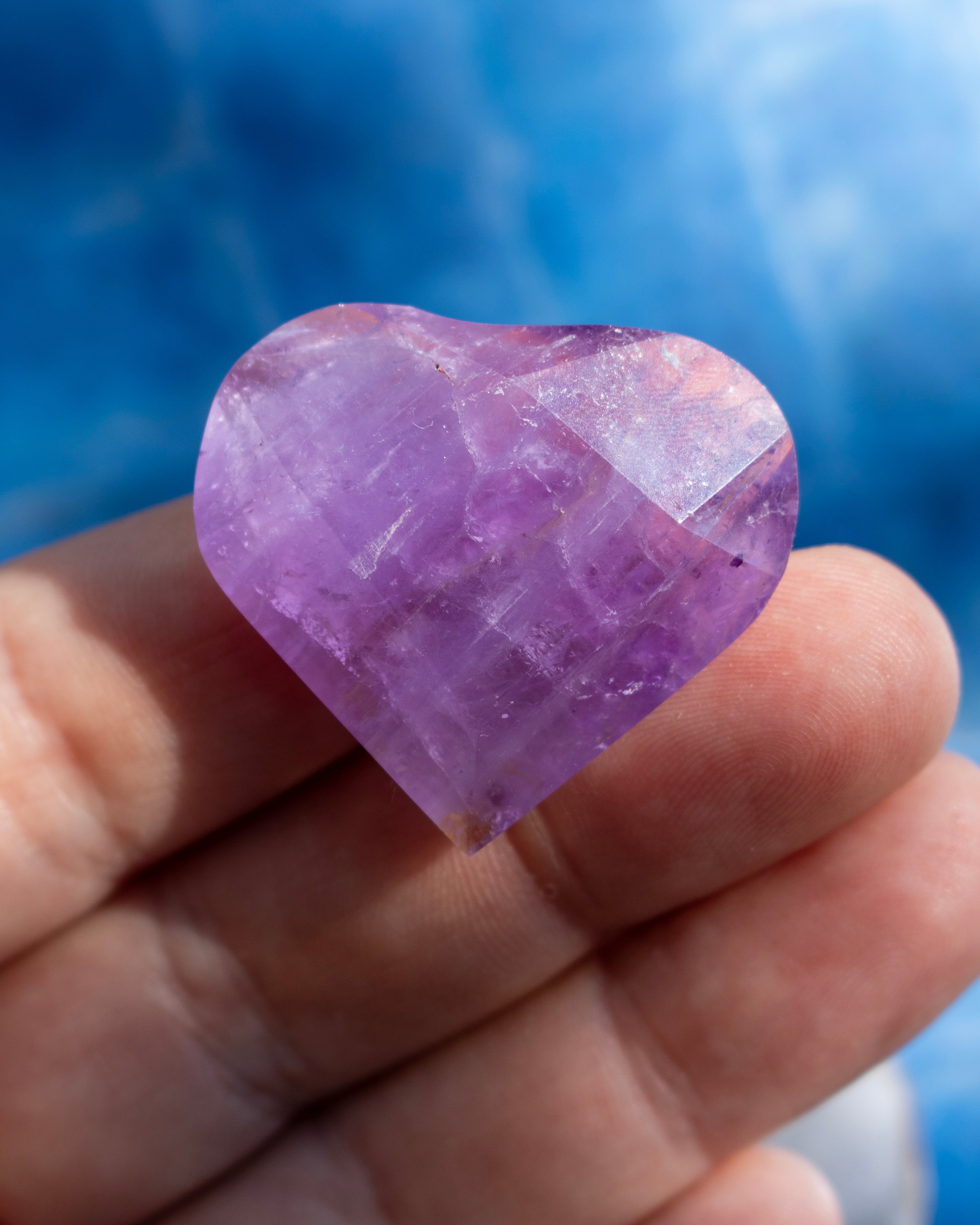 A heart-shaped purple amethyst gemstone held in a person's hand against a blurred blue background.