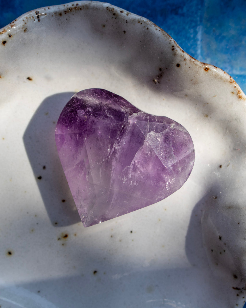 A heart-shaped purple amethyst crystal resting on a white ceramic dish with a blue surface visible in the background.