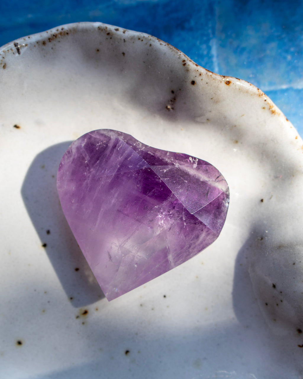 A heart-shaped purple amethyst crystal resting on a white ceramic dish with a blue textured background.