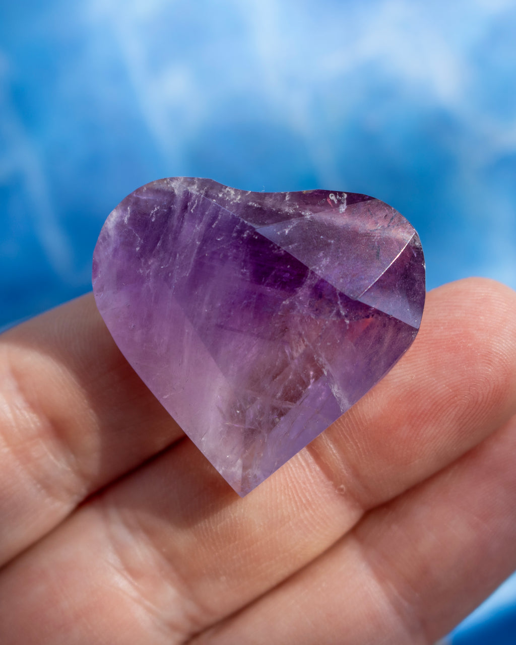 A heart-shaped purple amethyst gemstone held in a person's hand against a soft blue background.