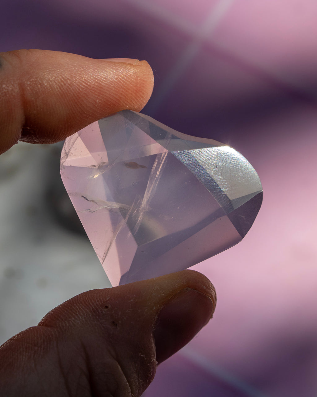 A faceted clear crystal gemstone held between two fingers, showing its translucent light purple-pink color and sharp geometric facets against a soft blurred purple background.
