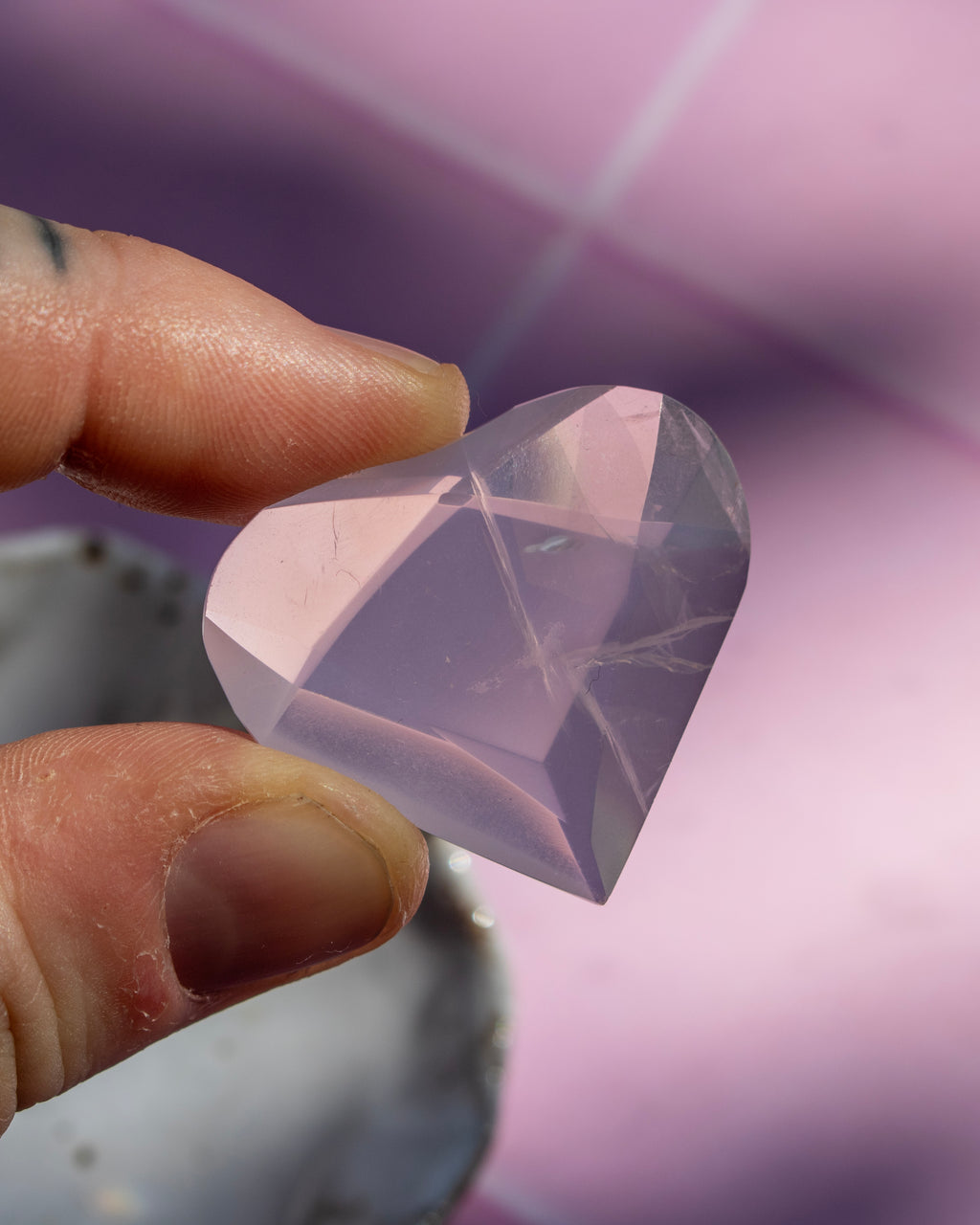 A faceted, heart-shaped clear crystal gemstone held between two fingers against a soft pink and purple blurred background.