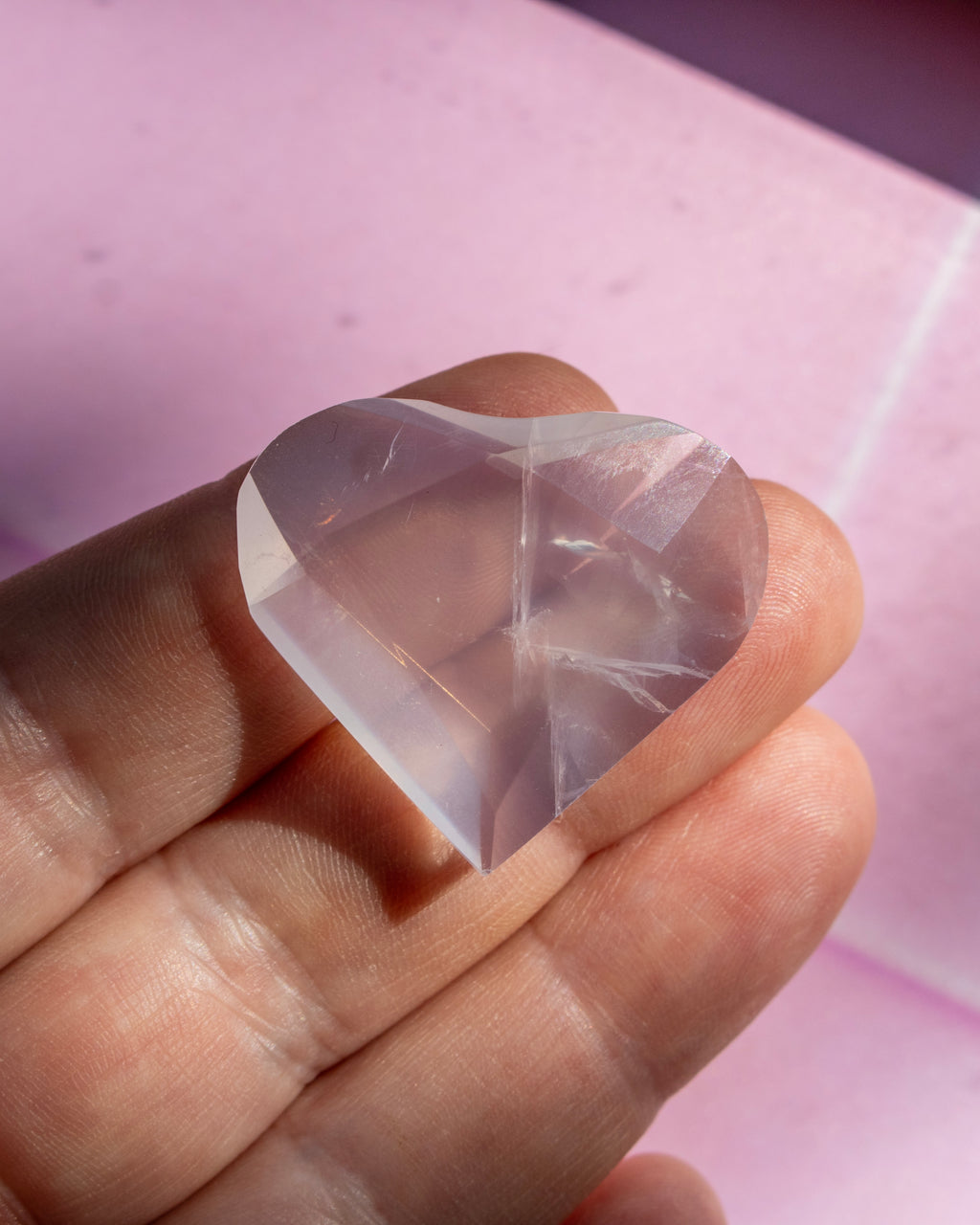 A faceted, heart-shaped clear crystal gemstone held between fingers against a soft pink background.