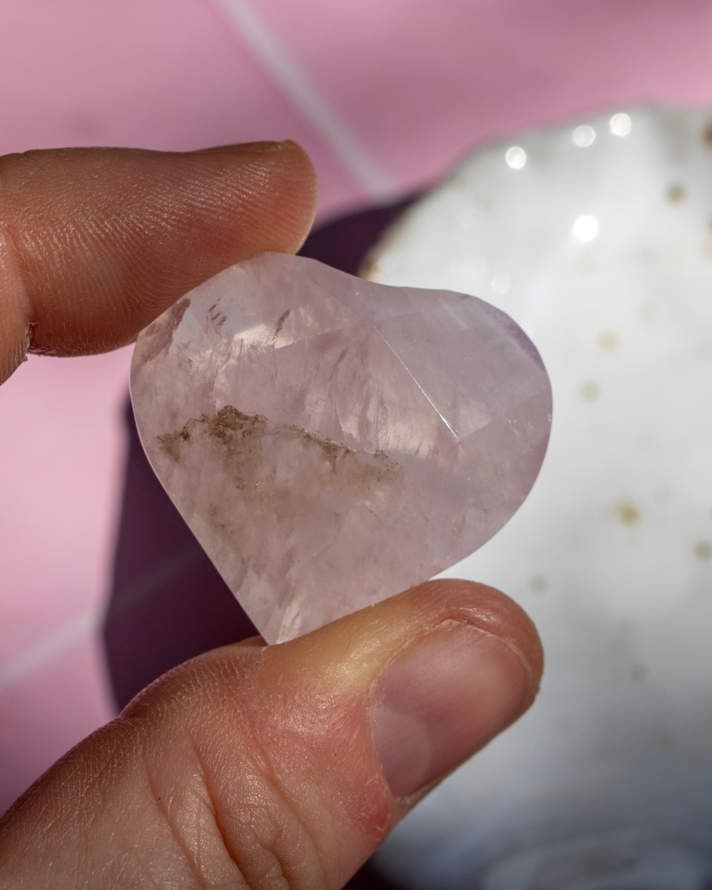 A heart-shaped pale pink crystal held between two fingers, with a blurred pink and white background.