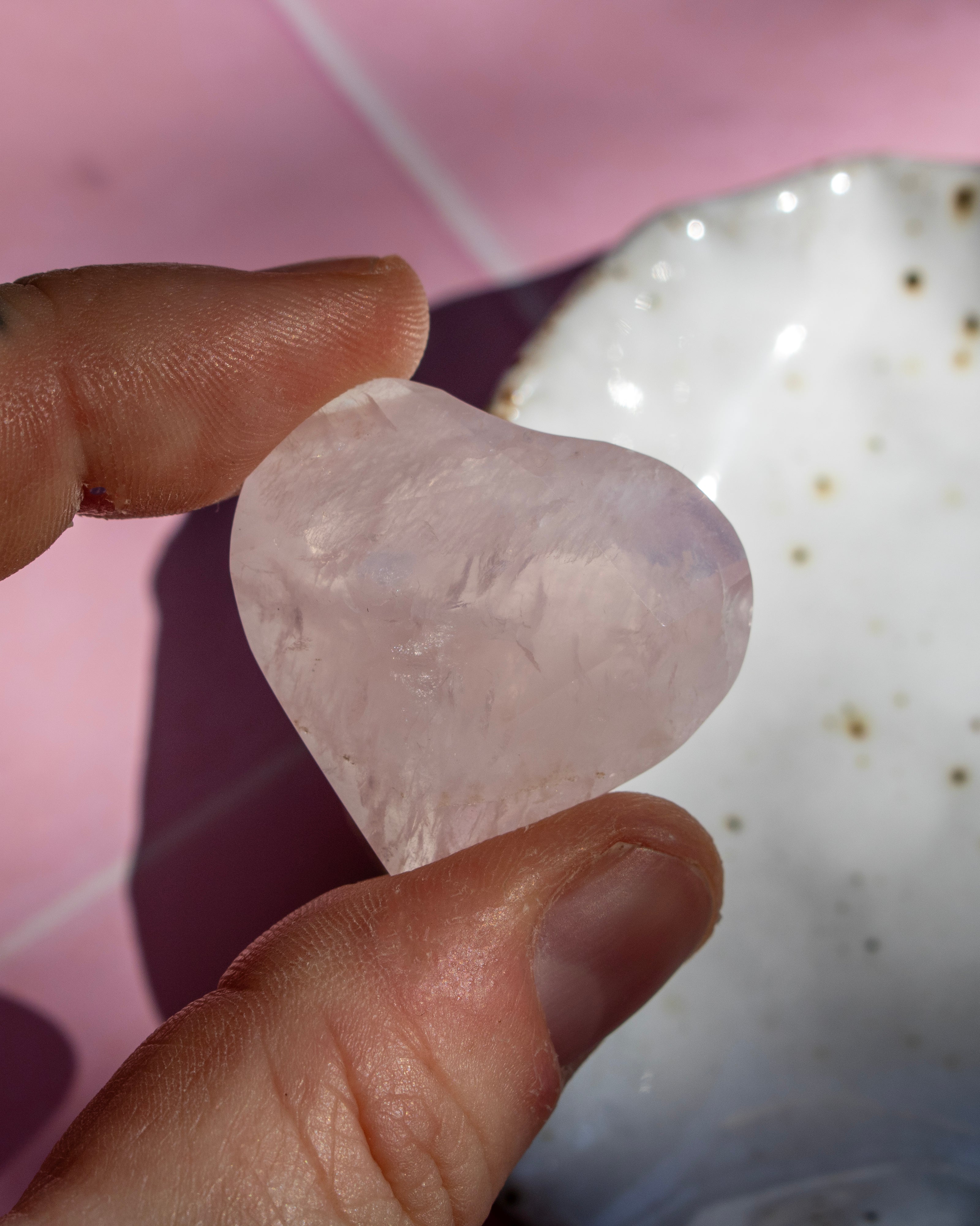A hand holding a polished, heart-shaped rose quartz crystal against a pink and white background.
