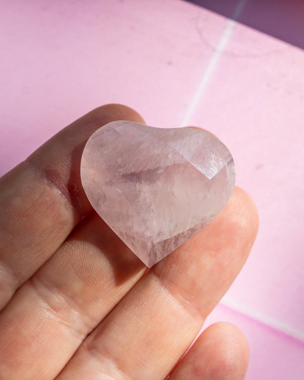 A faceted, heart-shaped clear crystal held on a person's fingers against a soft pink background.