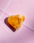 A heart-shaped translucent amber gemstone with natural inclusions, resting on a textured pink surface with a soft shadow.