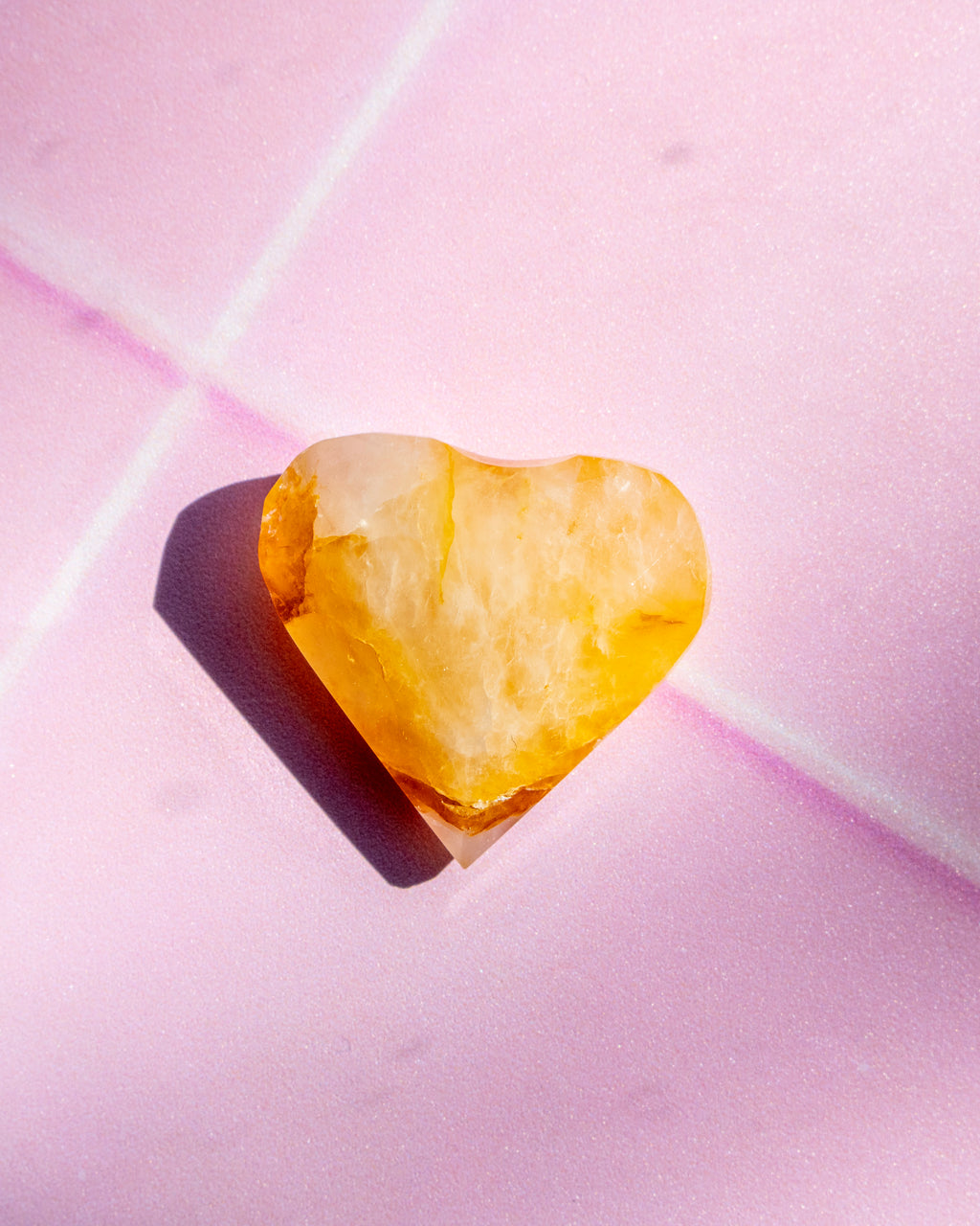 A small heart-shaped translucent yellow crystal gemstone casting a soft shadow on a textured light pink background.