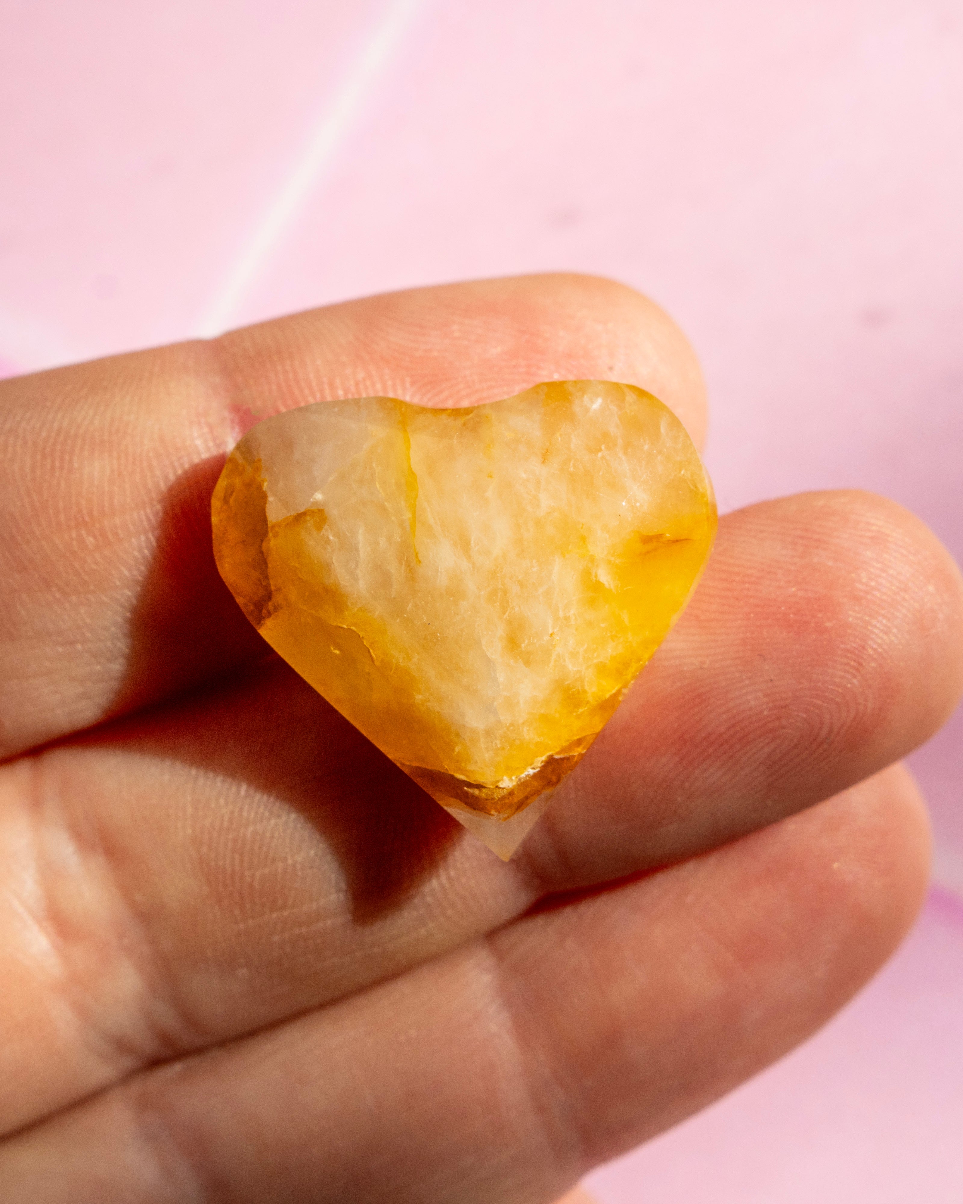 A heart-shaped raw crystal gemstone with a translucent yellow and cream color, held between fingers against a soft pink background.