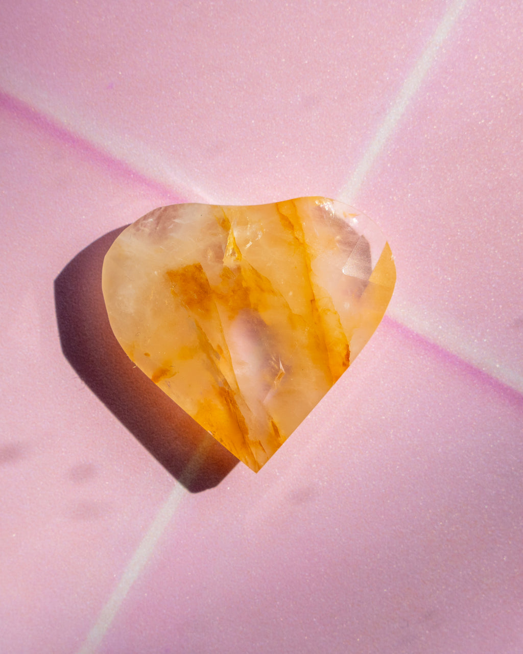 A heart-shaped translucent gemstone with warm amber and golden-brown inclusions, resting on a soft pink background with a cast shadow.