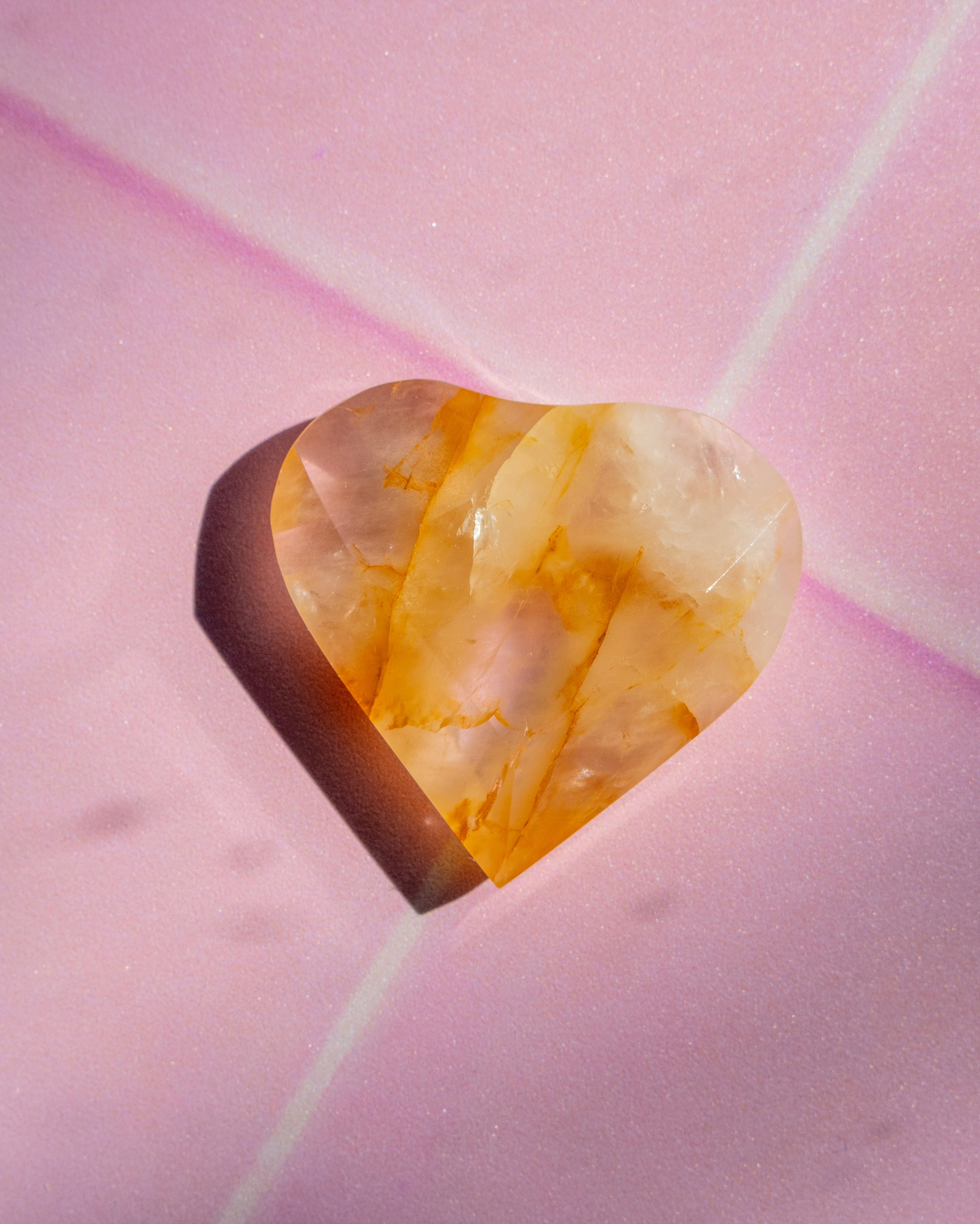 A heart-shaped faceted gemstone with marbled yellow and cream patterns, resting on a textured pink surface with a soft shadow.