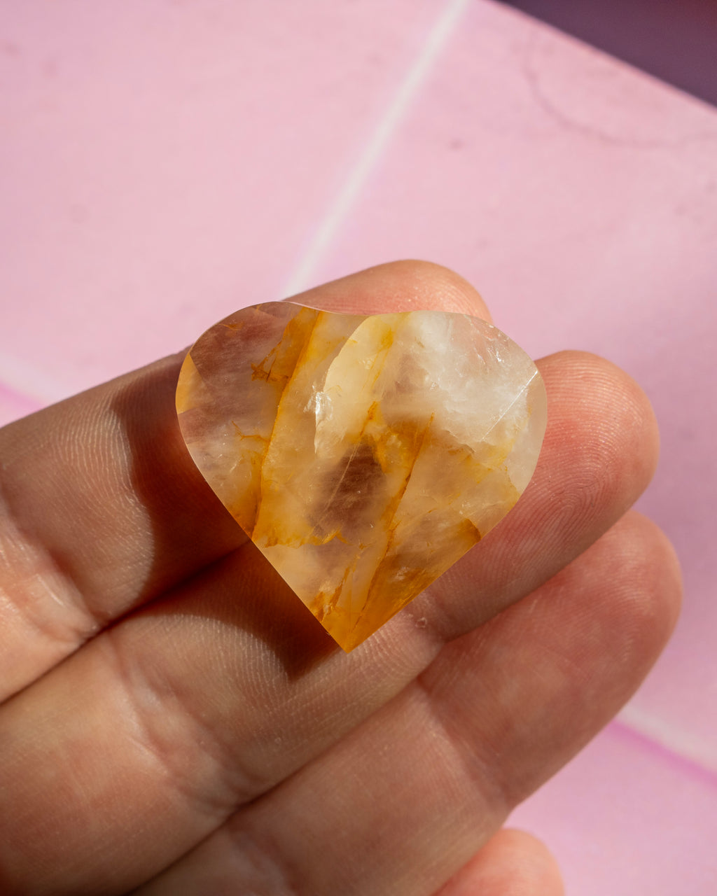 A heart-shaped raw crystal gemstone with natural orange and white banded patterns, held in a person's hand against a pink background.
