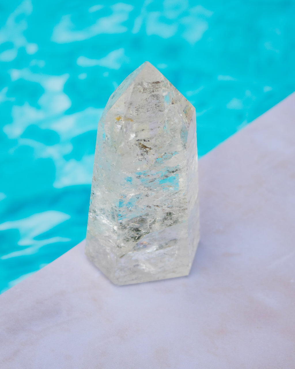A clear pointed crystal tower or point stone standing on a pale stone surface with bright turquoise water in the background.