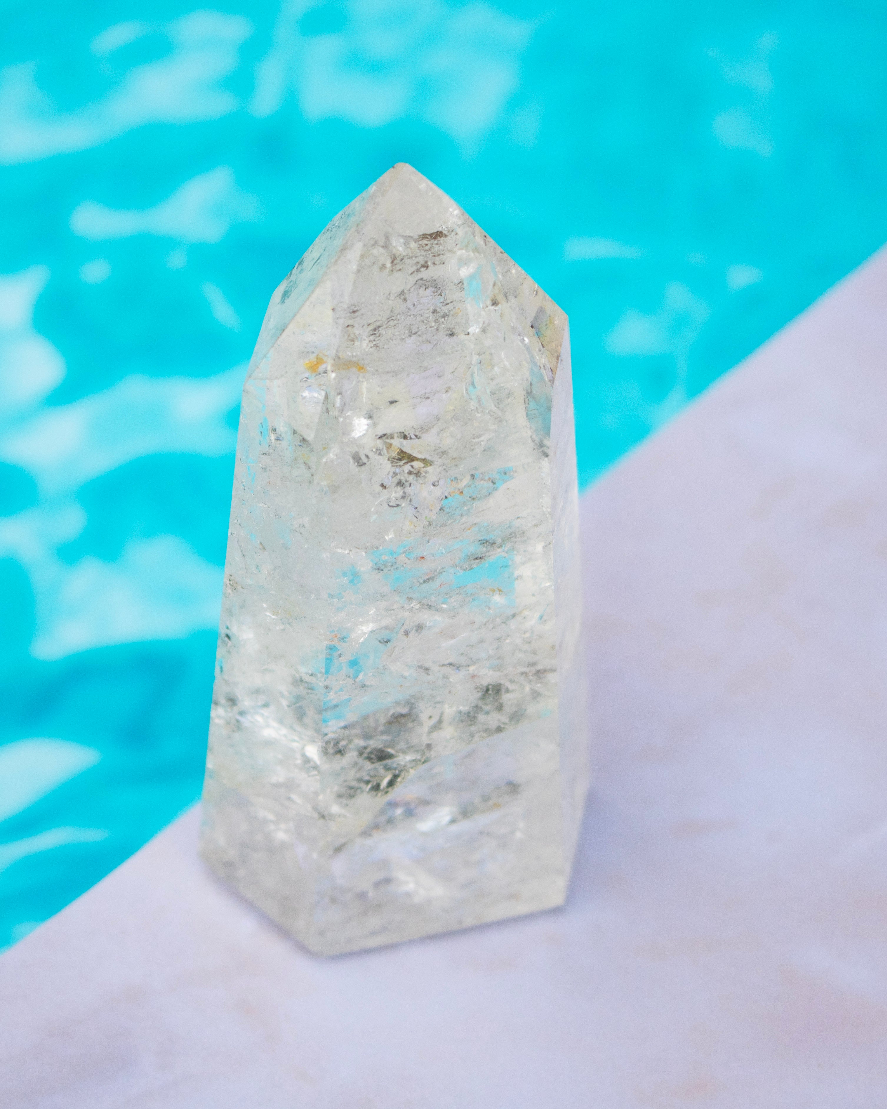 A clear quartz crystal point tower with natural inclusions, standing on a white surface with a bright turquoise blue background.