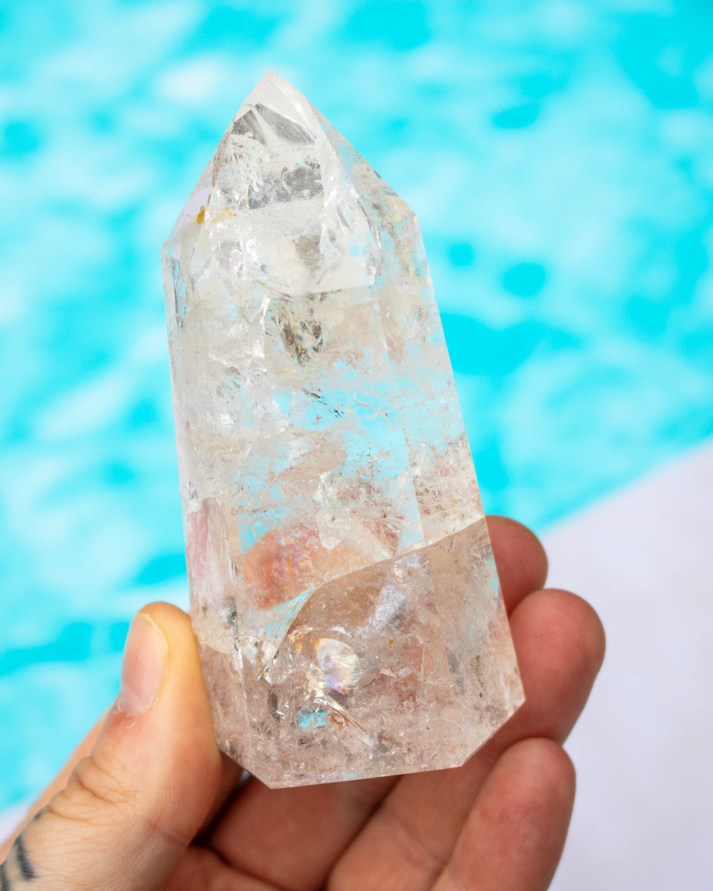 A hand holding a clear quartz crystal point with natural inclusions, set against a blurred turquoise blue background.